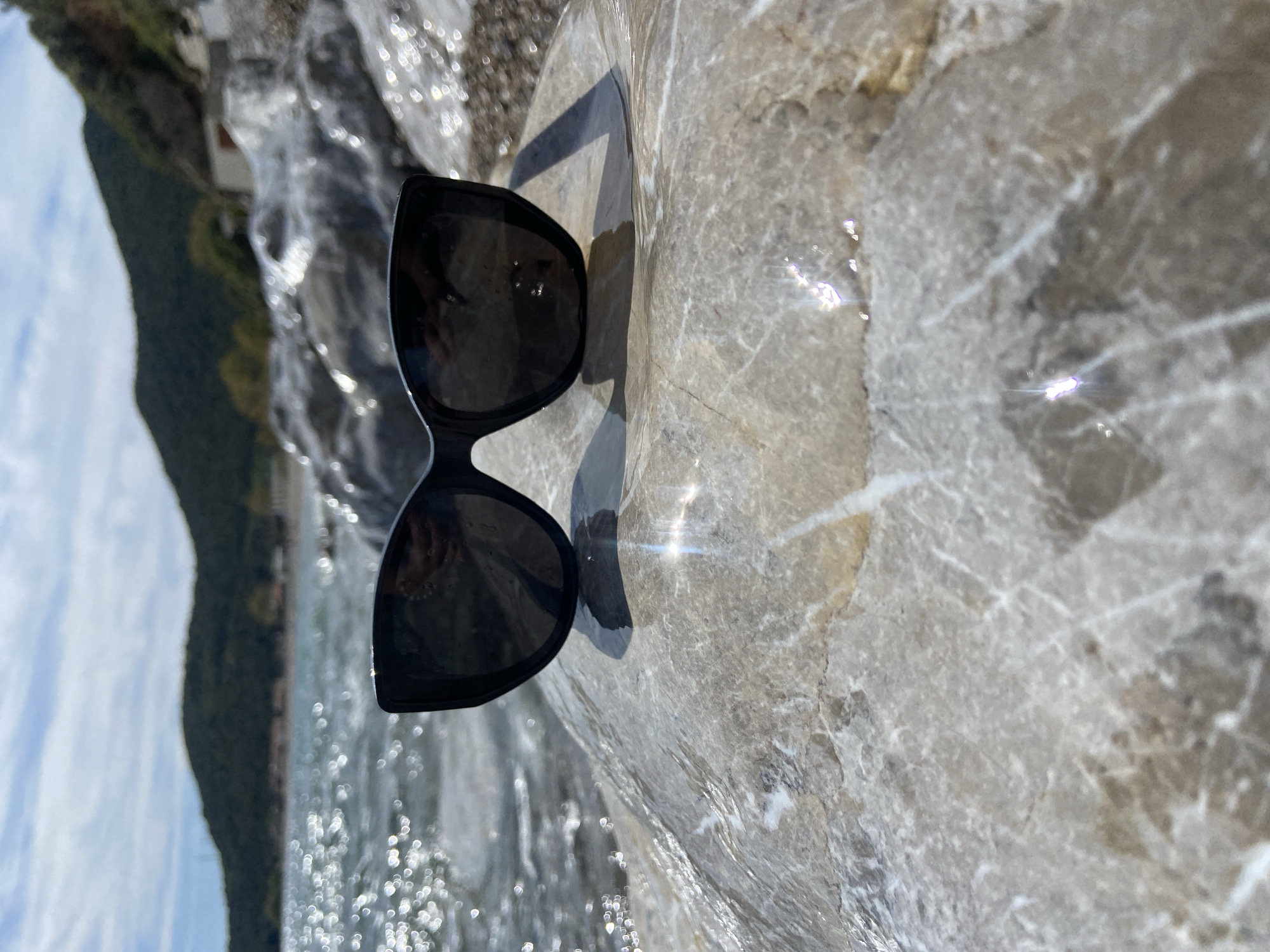The image shows a pair of black sunglasses lying on a light-colored, possibly marble, surface. The surface appears to be wet. In the background, slightly out of focus, there's a body of water, possibly a river, and a hillside with greenery. The location is Prijevor, Montenegro.