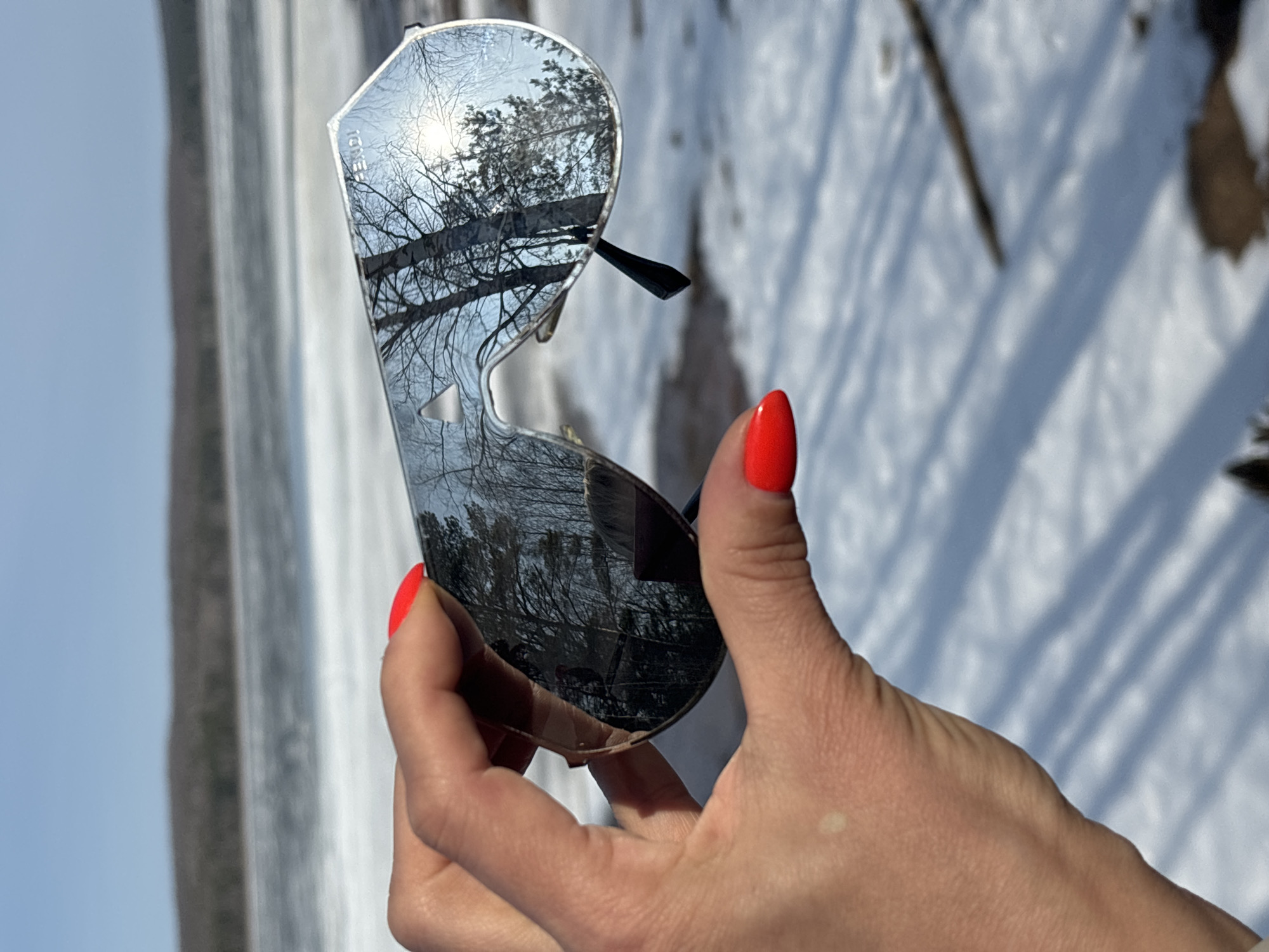 A person is holding a pair of silver sunglasses outdoors on a sunny day in what appears to be a winter scene, possibly near a snowy area. The sunglasses reflect the bare branches of trees and the bright sky. The person's hand is visible, with bright red nail polish. The background includes a possibly frozen body of water and the other side of its bank. In the far left of the image the gray surface of what appears to be a structure that would be next to that river-like body of water is visible. The location is implied to be Chaikovsky, Russia.