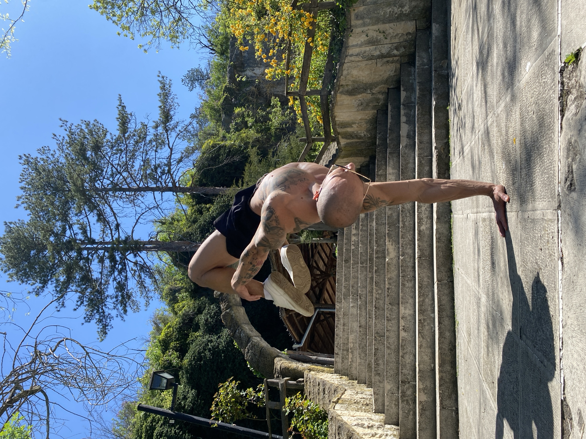 A shirtless man with dark shorts and white shoes is contorted in a yoga-like pose on stone steps, extending his body into a handstand with one hand on the step and the other holding his shoes, in Pleven, Bulgaria. The backdrop features lush green foliage, trees, and a clear blue sky. Stone stairs are also on the side with trees and greenery. A wooden structure is visible behind the man.