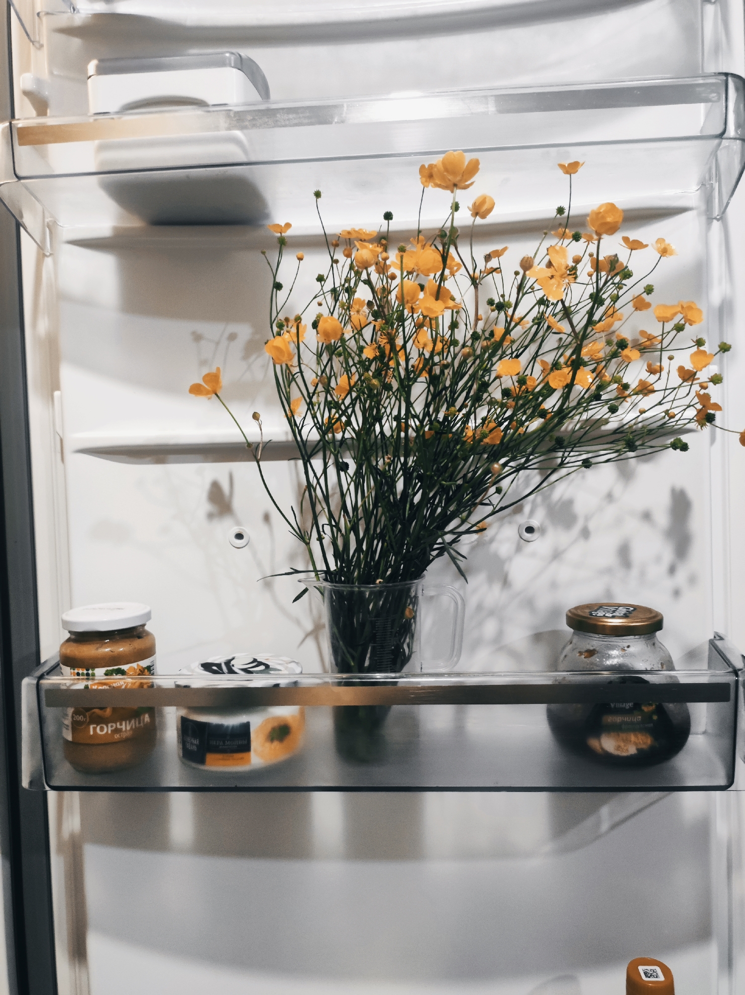 The image depicts the interior of a clean, white refrigerator in the city of Unknown, Unknown. A bouquet of vibrant yellow flowers, likely buttercups or ranunculus, with numerous green stems, is prominently displayed in a clear glass measuring cup, positioned on a transparent plastic shelf. The flowers cast soft shadows onto the white back wall of the refrigerator. Below the flowers, on a lower transparent door compartment, three items are stored. On the left, a glass jar with a white lid contains a light brown substance labeled "ГОРЧИЦА" (mustard). In the center, a small white and black container, possibly for dairy, features an orange and yellow design. To the right, a glass jar with a metallic gold lid holds a dark, viscous substance. The refrigerator walls are uniformly white, and the shelves are transparent plastic. A small white circular element is visible on the back wall of the refrigerator.