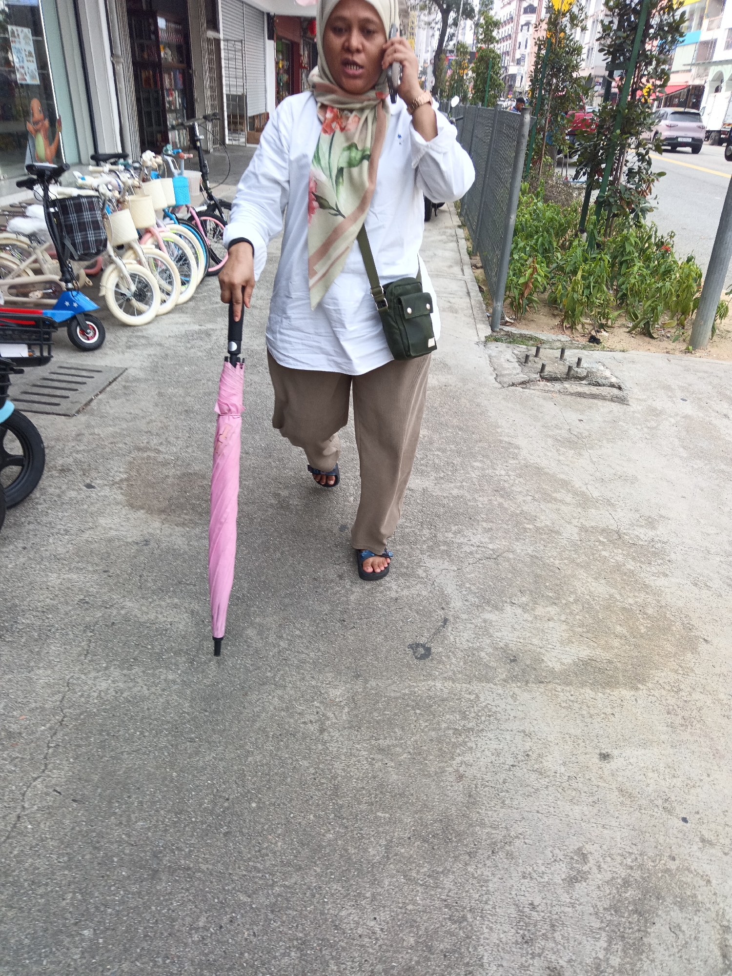 A woman wearing a hijab, white shirt, and brown pants walks along a sidewalk in Kuala Lumpur, Malaysia, holding a pink umbrella. She is talking on a mobile phone and carrying a dark green shoulder bag. In the background, several bicycles and an electric scooter are parked, and a street with cars and buildings is visible. The sidewalk is made of concrete and has a drain cover.