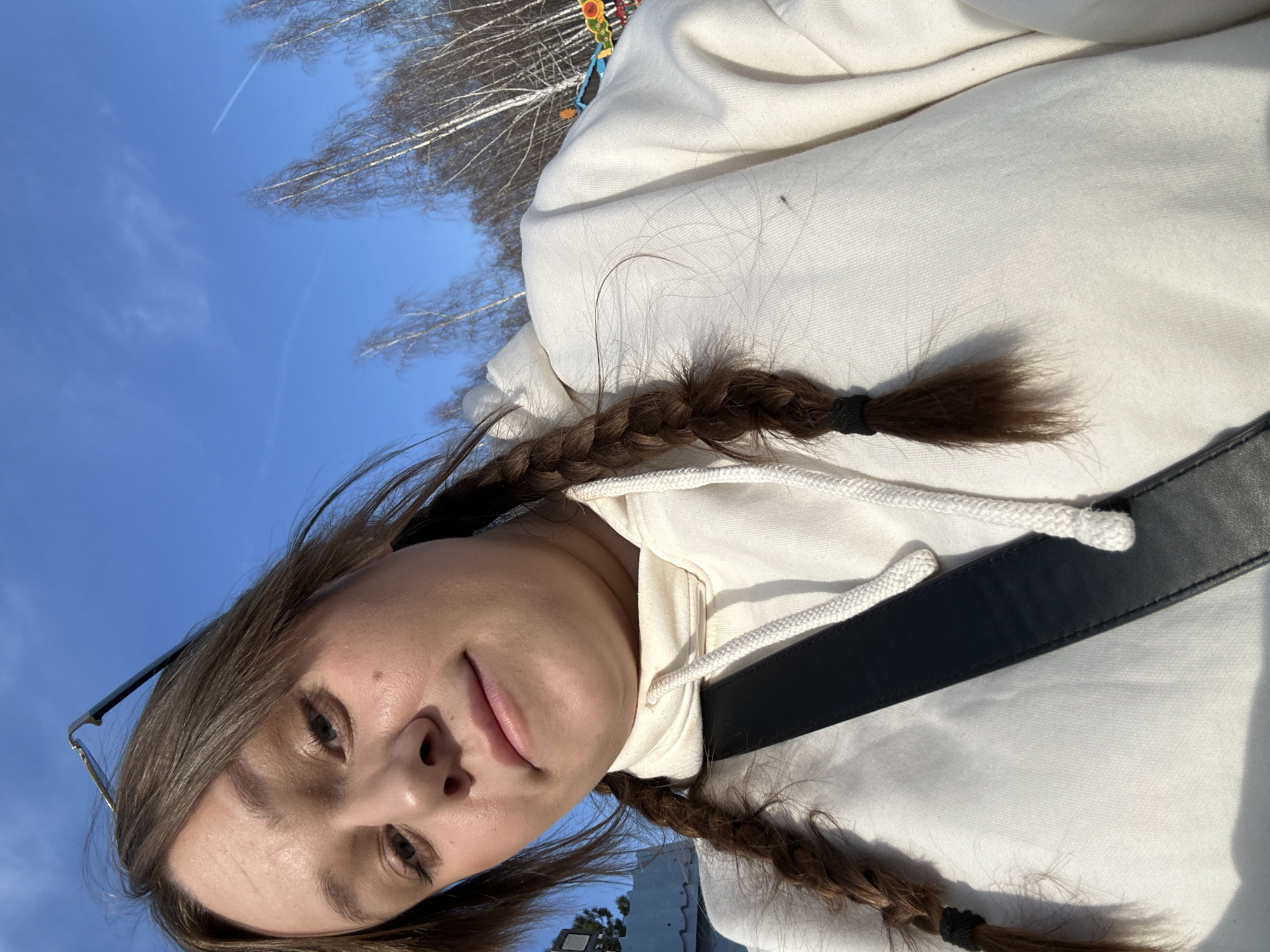 The image shows a young woman with brown hair styled in two braids, each secured with a black hair tie. She is wearing a white hoodie and a black shoulder strap is visible across her chest. She is outside, as evidenced by the clear blue sky and bare trees in the background. The photo appears to have been taken in Chaikovsky, Russia, as this was specified in the request.