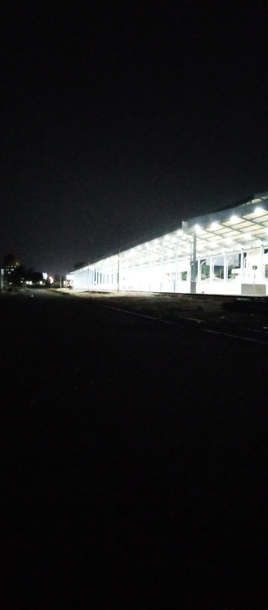 The image captures a scene at night, likely a railway station, in Kedungrejo, Indonesia. A long, covered structure, presumably a platform or waiting area, is brightly illuminated by overhead lights. The structure extends diagonally across the frame, with its roof supported by numerous evenly spaced columns. The area in front of the structure appears to be a track or pathway, with some low vegetation visible alongside it. In the distance, other lights suggest the presence of more buildings or infrastructure within the town. The sky is dark and overcast, typical of nighttime. The overall impression is of a functional, well-lit infrastructure facility in a rural or semi-urban Indonesian setting.