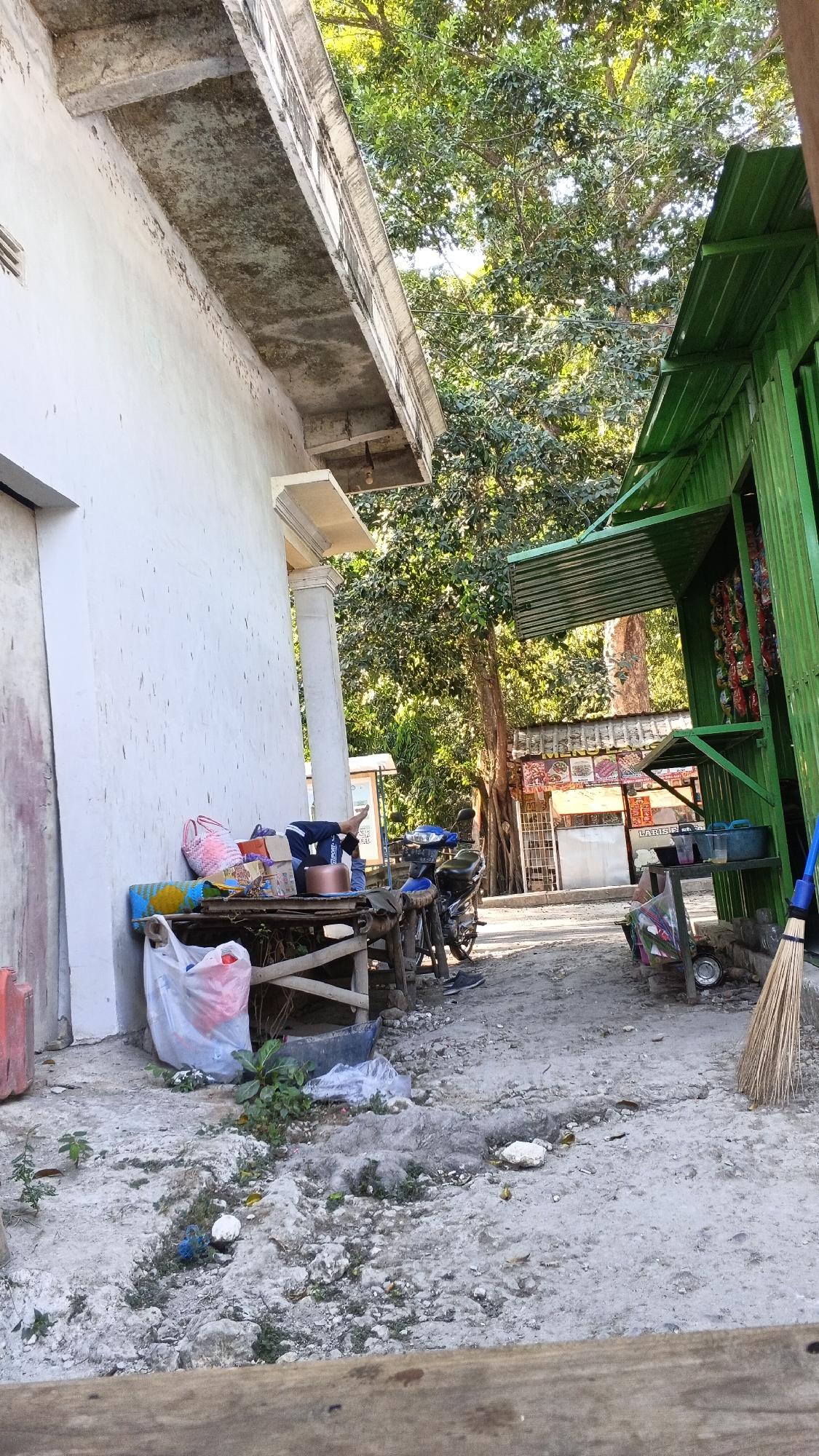 The image depicts an outdoor scene in Rengel, Indonesia, likely a roadside area. A white building with peeling paint is on the left, with a concrete overhang. In the center, a makeshift wooden table displays various items including bags, boxes, and a metallic pot. A person, partially visible, is resting on the ground next to the table with their legs extended. A motorcycle is parked nearby. To the right, a green stall with a corrugated metal roof displays goods, possibly snacks or small items. A broom stands against the stall. The ground is uneven and dusty, with some scattered debris and vegetation. In the background, a large tree with dense foliage dominates the scene, with other buildings and signs partially visible further down the road. The overall impression is of a simple, possibly commercial, roadside environment.