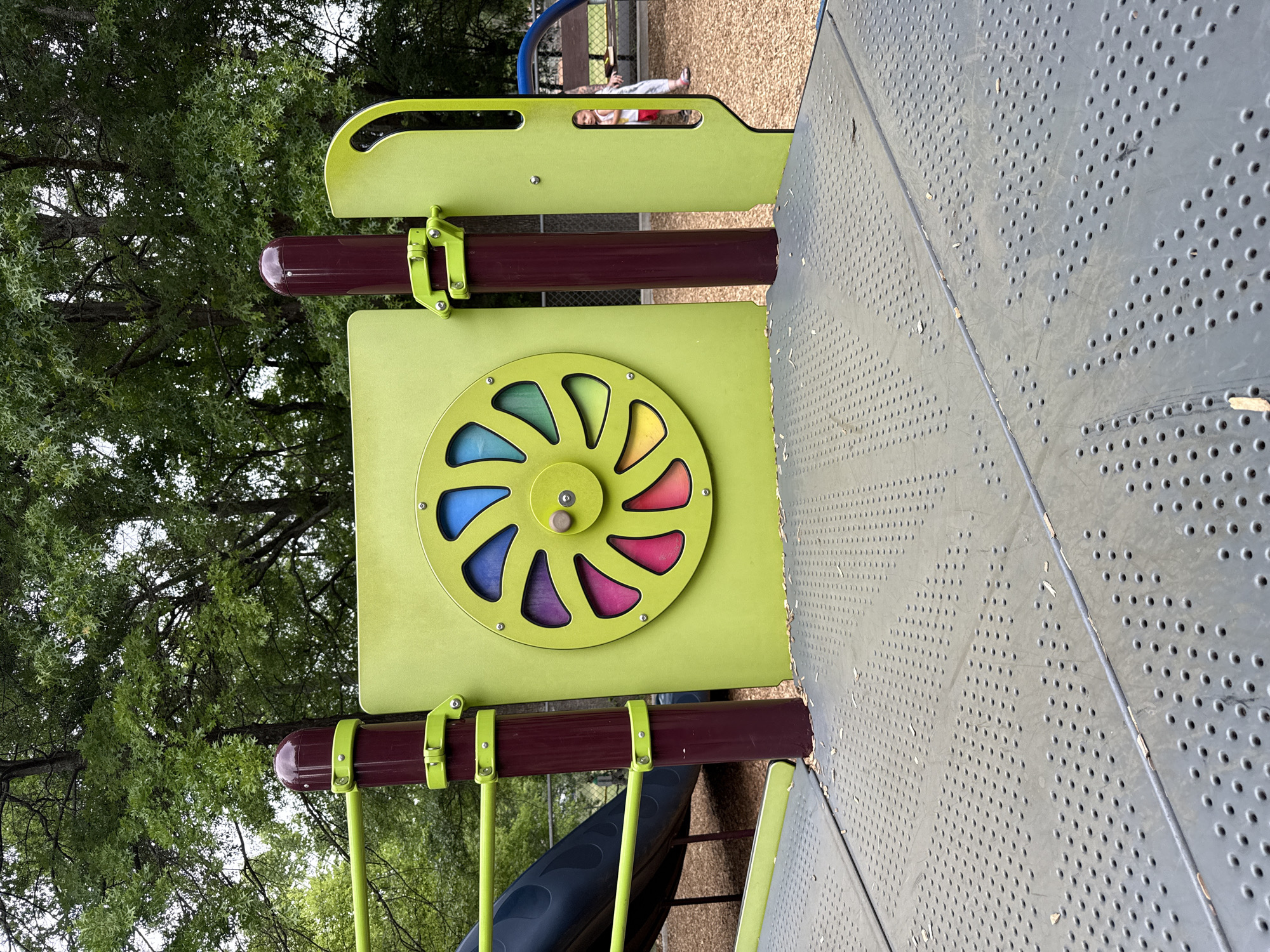 The image shows a section of a playground structure, likely in Mountain Park, United States of America. A green panel with a circular, multicolored wheel featuring sections in blue, purple, yellow, and red is prominently displayed. This panel is connected to two dark brown posts with light green clamps. A gray metal platform with small holes is adjacent to the green panel. In the background, trees are visible. The overall setting suggests a park or recreational area. A child is visible in the background.