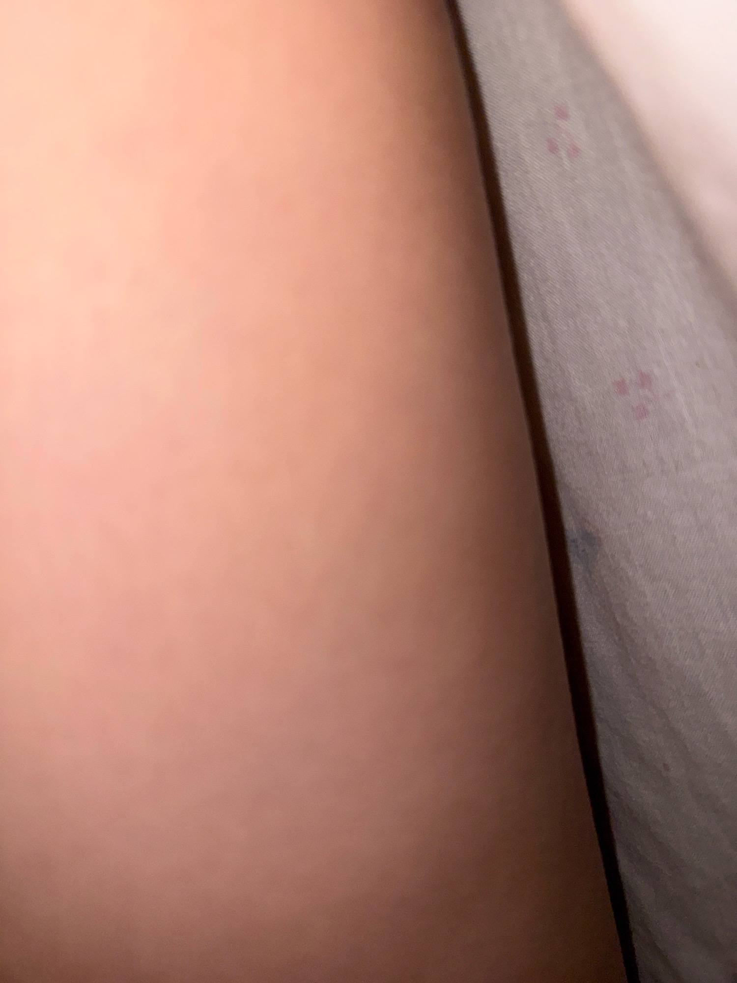 The image shows a close-up, low-angle view of a person's skin, appearing to be an arm or leg, against a light-colored fabric with a subtle pattern of pinkish dots. The skin is smooth and shows subtle variations in tone, with a warm, peachy hue. The fabric is creased and folded, suggesting it might be bedding or clothing. The lighting is soft, casting gentle shadows and highlighting the textures of both the skin and the fabric. The image is abstract, focusing on the tactile and visual qualities of the materials rather than a specific scene or location. There are no discernible elements that would allow for the identification of a specific location in Zavolzhye, Russia.
