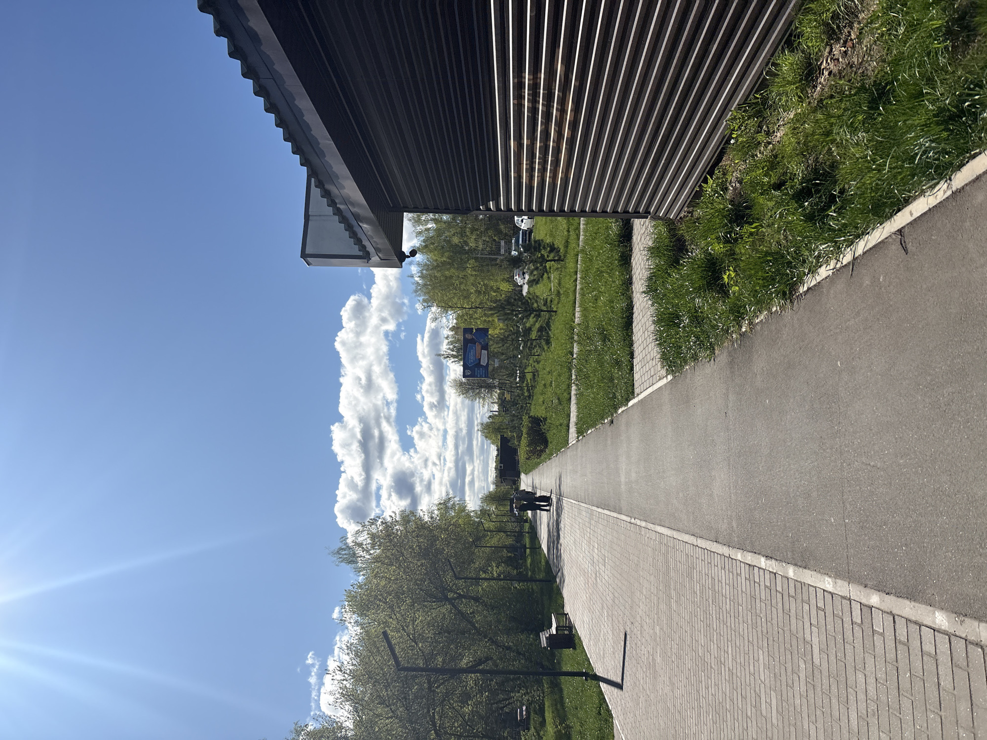 A horizontal, outdoor shot depicts a sidewalk and surrounding areas in the city of Chaikovsky, Russia. The sidewalk, constructed from light gray paving stones on one side and dark gray asphalt on the other, stretches into the distance. To the left of the sidewalk, trees line the path, casting long shadows on the walkway. Occasional benches are placed under the trees. To the right, short green vegetation grows alongside the sidewalk, bordering a building with corrugated metal walls. The sky is blue with fluffy white clouds. In the distance, a billboard is visible. Two people stand farther down the sidewalk.