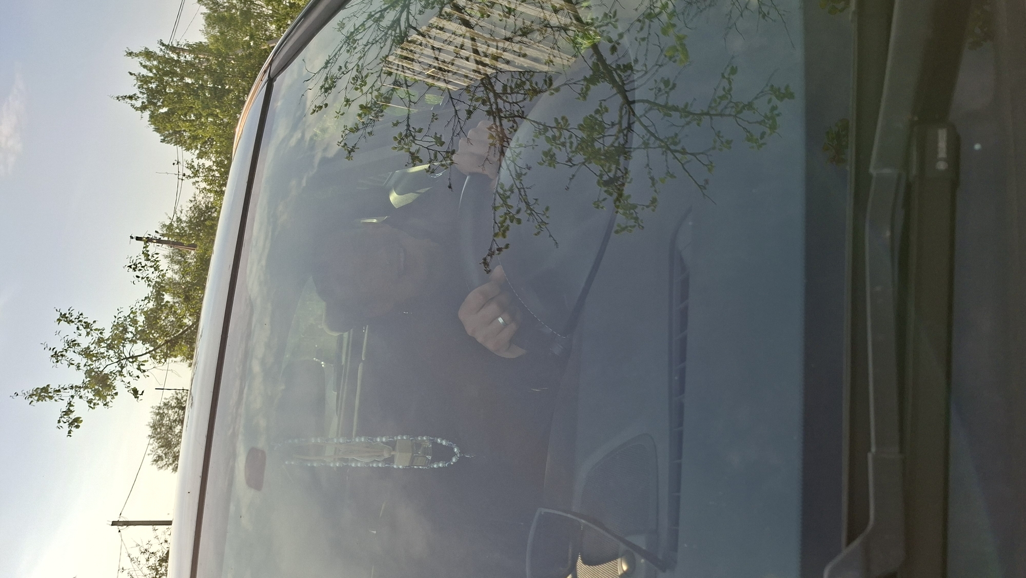 A person, possibly female, is seen driving a car. They are wearing dark clothing and a ring on their left hand. The person's face is partially obscured by glare and shadow, but they appear to be smiling. A string of beads with a religious charm hangs from the rearview mirror. The car's windshield reflects trees and a building. The scene appears to be set in a rural or suburban area, possibly within the Забалоцкі сельскі Савет in Belarus, given the presence of trees, utility poles, and a cloudy sky suggestive of daytime.