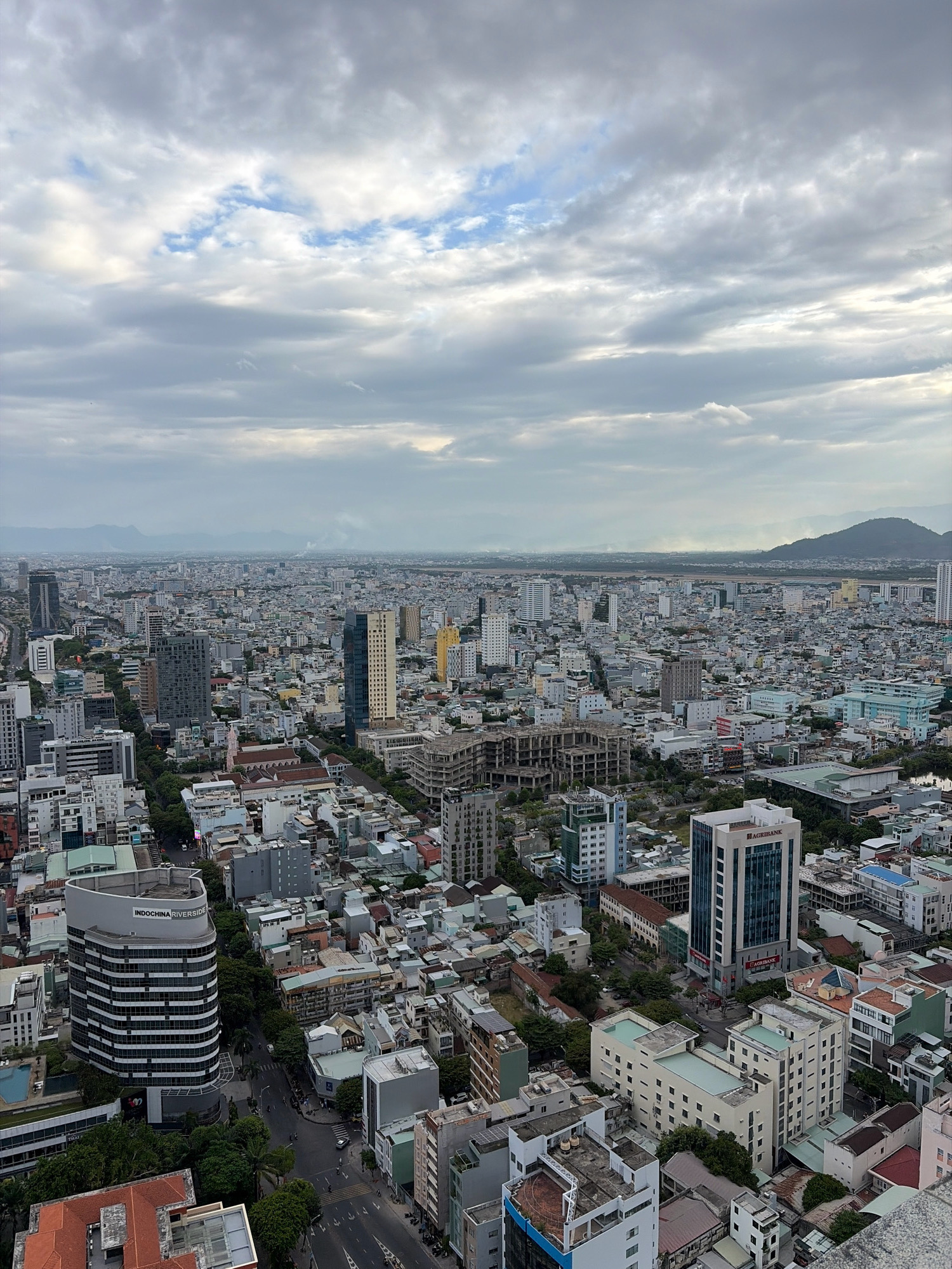 This is an aerial, wide-angle photograph of the city of Da Nang, Vietnam, under a cloudy sky. The city features a dense urban landscape with numerous multi-story buildings of varying architectural styles and heights, ranging from mid-rise apartments to taller office buildings. Trees are interspersed throughout the cityscape, particularly along streets and within some open courtyards. The buildings are predominantly light-colored, with some featuring glass facades and balconies. In the foreground, a curved, modern office building with the text "INDOCHINA RIVERSIDE" is visible. Further into the distance, rows of smaller buildings give way to a more sprawling urban expanse. Mountains are visible on the horizon, shrouded in a light haze. The lighting suggests it is either early morning or late afternoon, with diffuse light filtering through the clouds. No individuals are clearly discernible in the image, and no specific actions or interactions are apparent. The scene depicts a typical urban environment in a coastal Southeast Asian city.