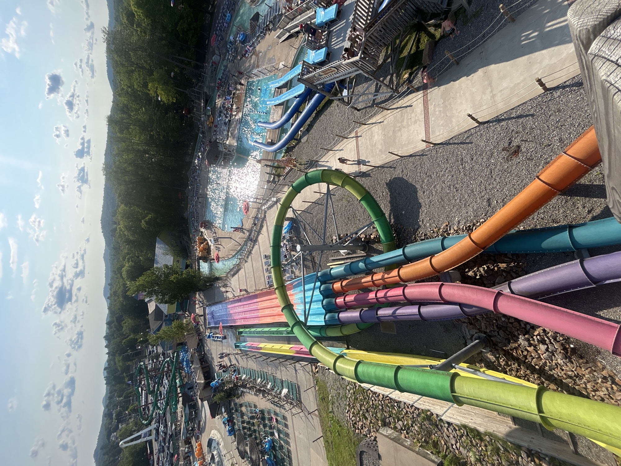 The image shows a view from above of a water park in Old Forge, New York. In the foreground, a multi-colored water slide descends, with tubes in green, yellow, pink, purple, orange, and blue. To the right, a section of a swimming pool is visible, with lounge chairs and umbrellas surrounding it, and people swimming. Further back, more water slides, including a large green spiral slide, are visible, along with a parking lot filled with cars. The park is set against a backdrop of lush green trees and hills under a bright blue sky with scattered clouds.