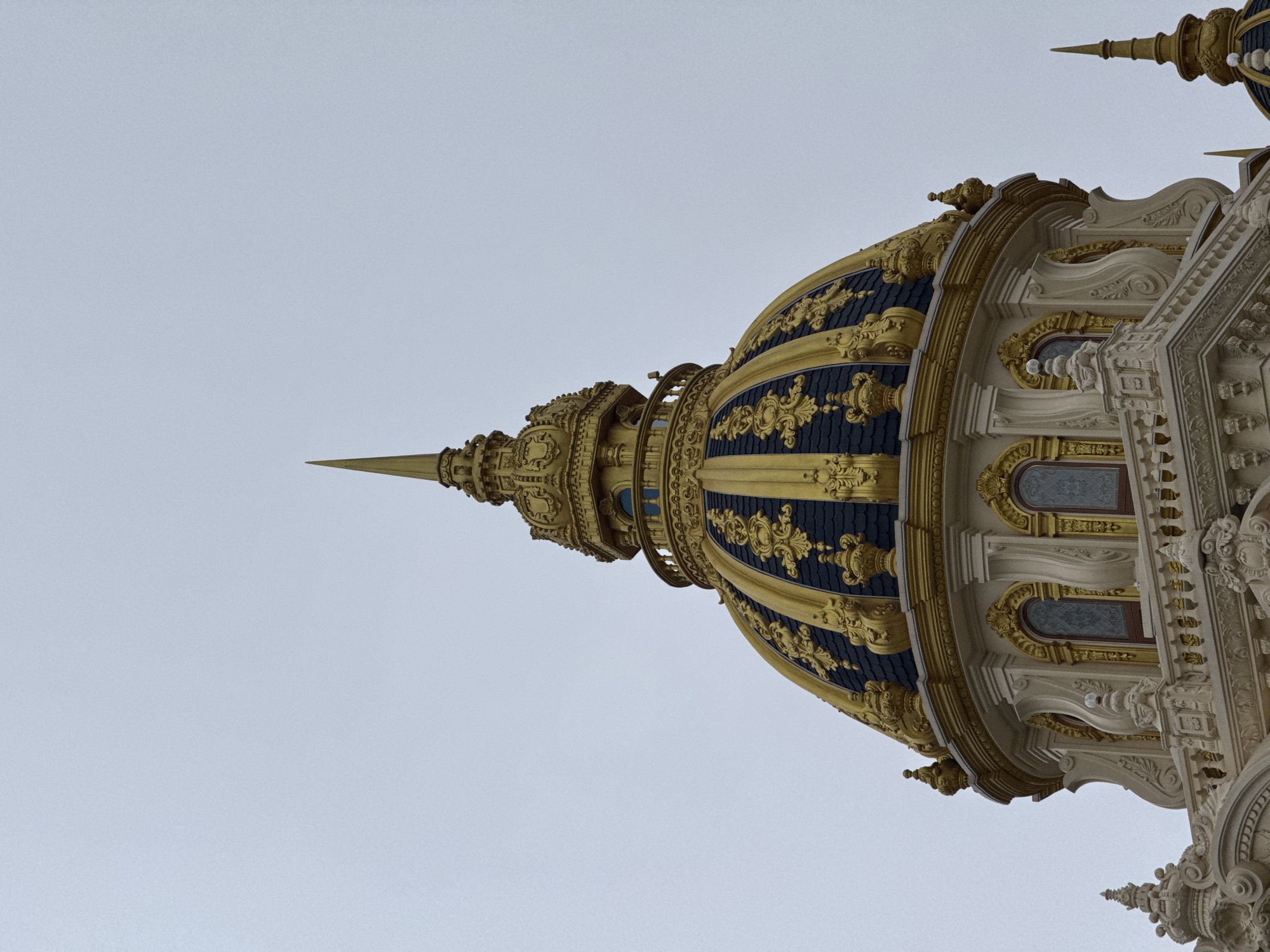 The image shows a close-up view of a golden dome atop a building with white architectural details and ornate gold trim. The dome is dark blue with intricate gold embellishments and a golden spire at its apex. Several arched windows with stained glass are visible below the dome, surrounded by white, sculpted structures. The sky is overcast and pale gray. The building is the Dinh Bao Dai, also known as the Summer Palace of Emperor Bao Dai, located in Da Lat, Vietnam.