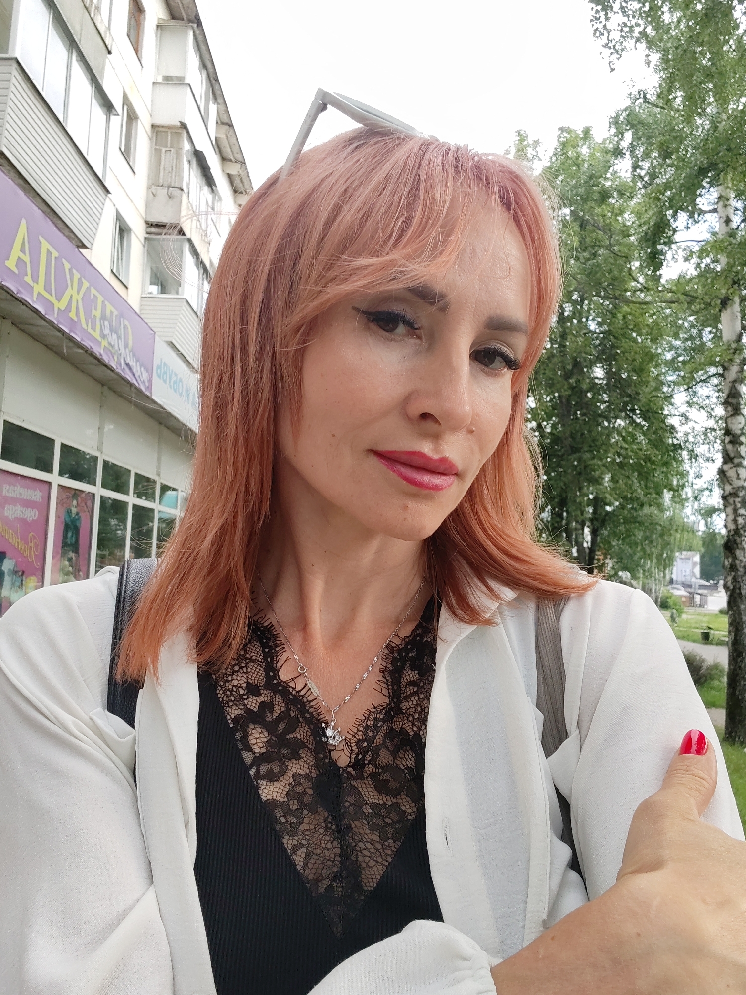 A selfie shows a woman with reddish-pink hair and heavy makeup posing outdoors. She is wearing a white open shirt over a black lace top, and a silver necklace. Her right hand is visible in the foreground, with red nail polish. In the background, there is a building with balconies and windows, and trees. A purple sign is visible on the building, with Russian text that appears to read "АПЖЕРА" (APZHERA). This type of building and sign is consistent with urban settings in Russian cities, including Chaikovsky, Russia.