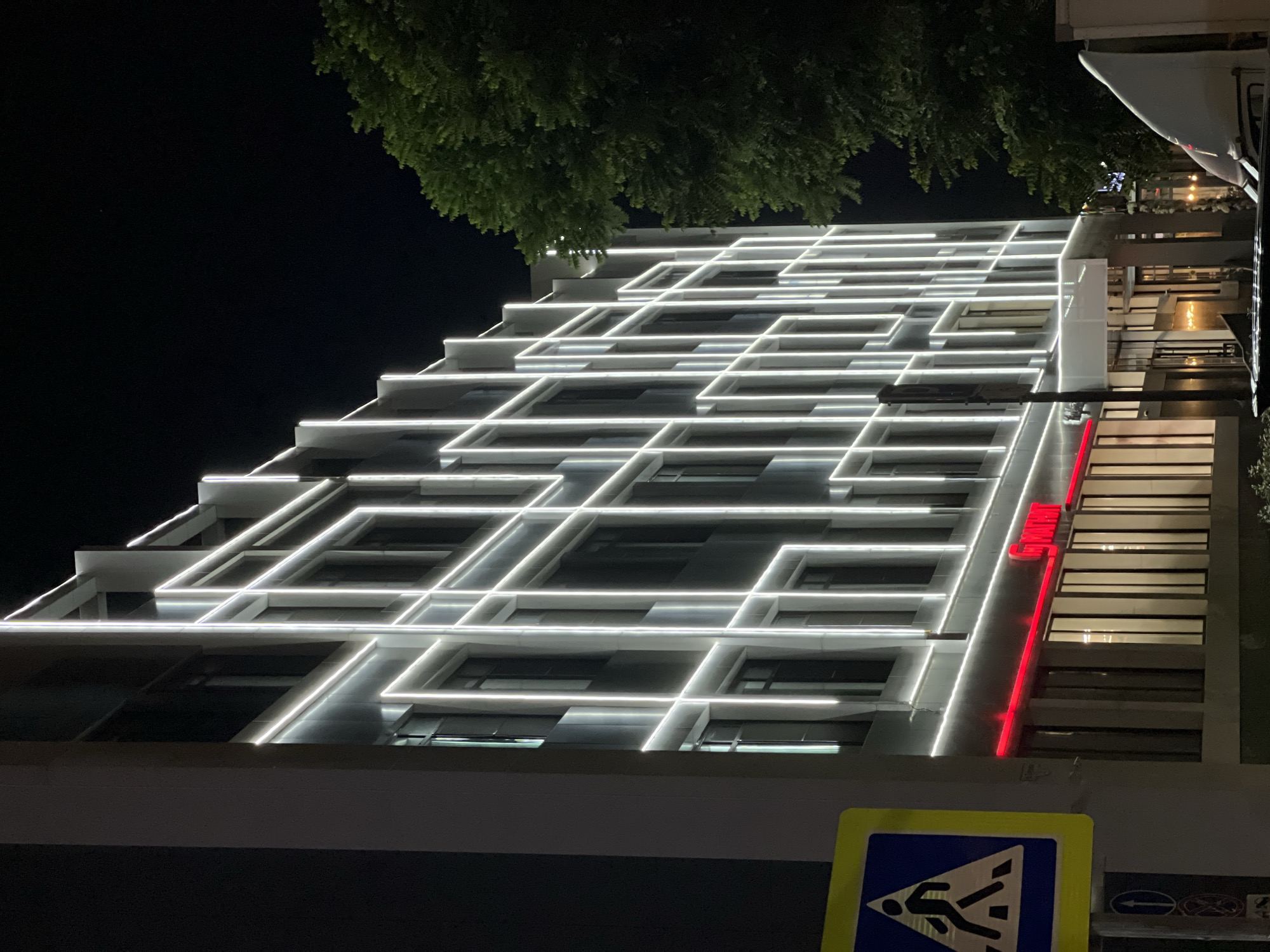A multi-story building with a modern facade illuminated by white LED lights is captured at night in Krasnodar, Russia. The building features a grid-like pattern of lights outlining its geometric structure, creating a striking visual effect against the dark sky. Red neon signage is visible on the lower level of the building, suggesting a commercial establishment. A pedestrian crossing sign with a yellow border and a blue interior depicting a person walking is prominently displayed on the left side of the image. A large, dark green tree is situated to the right of the building, its foliage contrasting with the man-made illumination. The overall scene depicts an urban environment at night, with the architecture and lighting being the central focus.