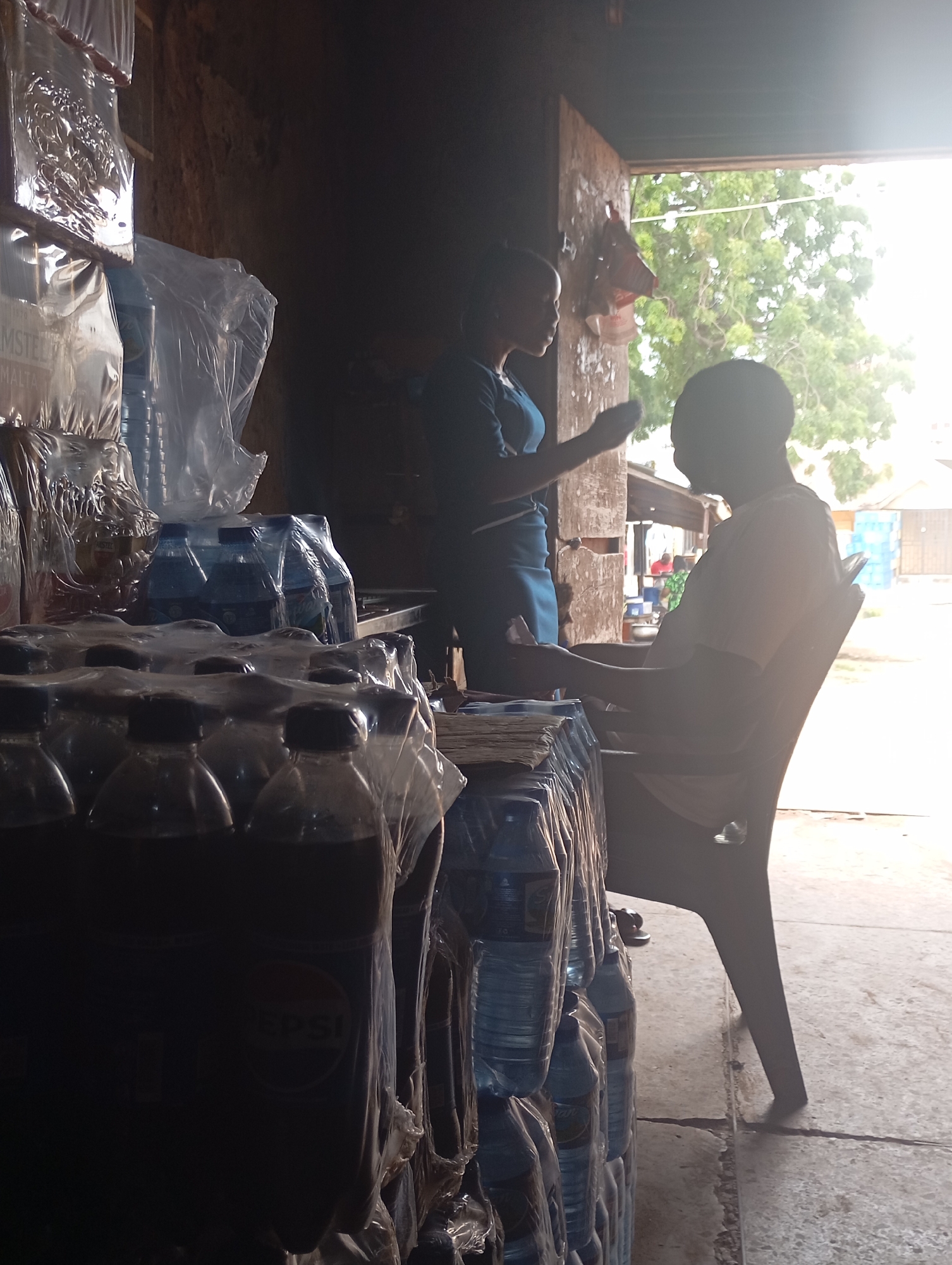 In Shendam, Nigeria, a woman in a dark blue dress stands behind a counter laden with bottled drinks, her left hand extended. Facing her, a person in a light-colored t-shirt sits in a dark plastic chair, their right arm resting on the armrest. They are engaged in what appears to be a transaction. The environment is dim, with bright light filtering in from an open doorway in the background, illuminating trees and buildings. The foreground is dominated by stacks of plastic-wrapped bottled drinks, including visible Pepsi branding on some. The floor is concrete, marked by cracks.