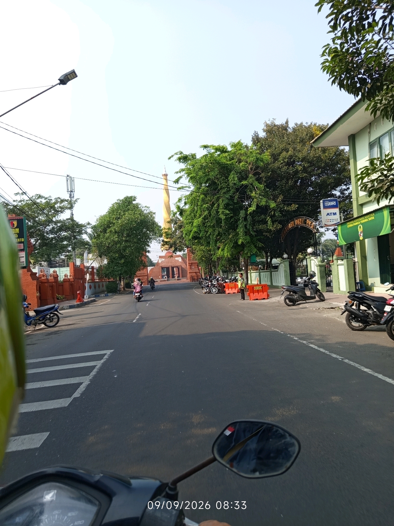 The image depicts a street scene in Mojokerto, Indonesia, likely near the Trowulan archaeological site given the presence of a large, golden stupa-like monument in the background, which is reminiscent of structures found there. The foreground shows a portion of a motorcycle from a first-person perspective, with the handlebars and part of the dashboard visible. The road is paved and has white lane markings. On the left side of the road, there is a blue motorcycle parked, and further down, a red brick wall with intricate carvings and decorative elements lines the street. A tall, modern street lamp with LED lights is also visible in the upper left portion of the image.
In the middle ground, there are several people on motorcycles, some of whom are wearing headscarves. The golden monument, the focal point of the background, is a towering structure with a distinct shape, possibly a reliquary or a replica of an ancient religious monument. To the right of the road, a light green building with windows and a sloped roof stands, with trees and foliage surrounding it. A sign on the building indicates "BRI ATM" and another green banner displays text in Indonesian, likely advertising a business or cooperative. The sky is clear and bright, suggesting it is daytime. Orange traffic cones are placed along the sidewalk, and several parked motorcycles are visible in front of the building. A security guard or attendant in a yellow vest stands near the cones. The overall impression is a bustling urban street in Indonesia, with a blend of modern infrastructure, historical elements, and daily life.