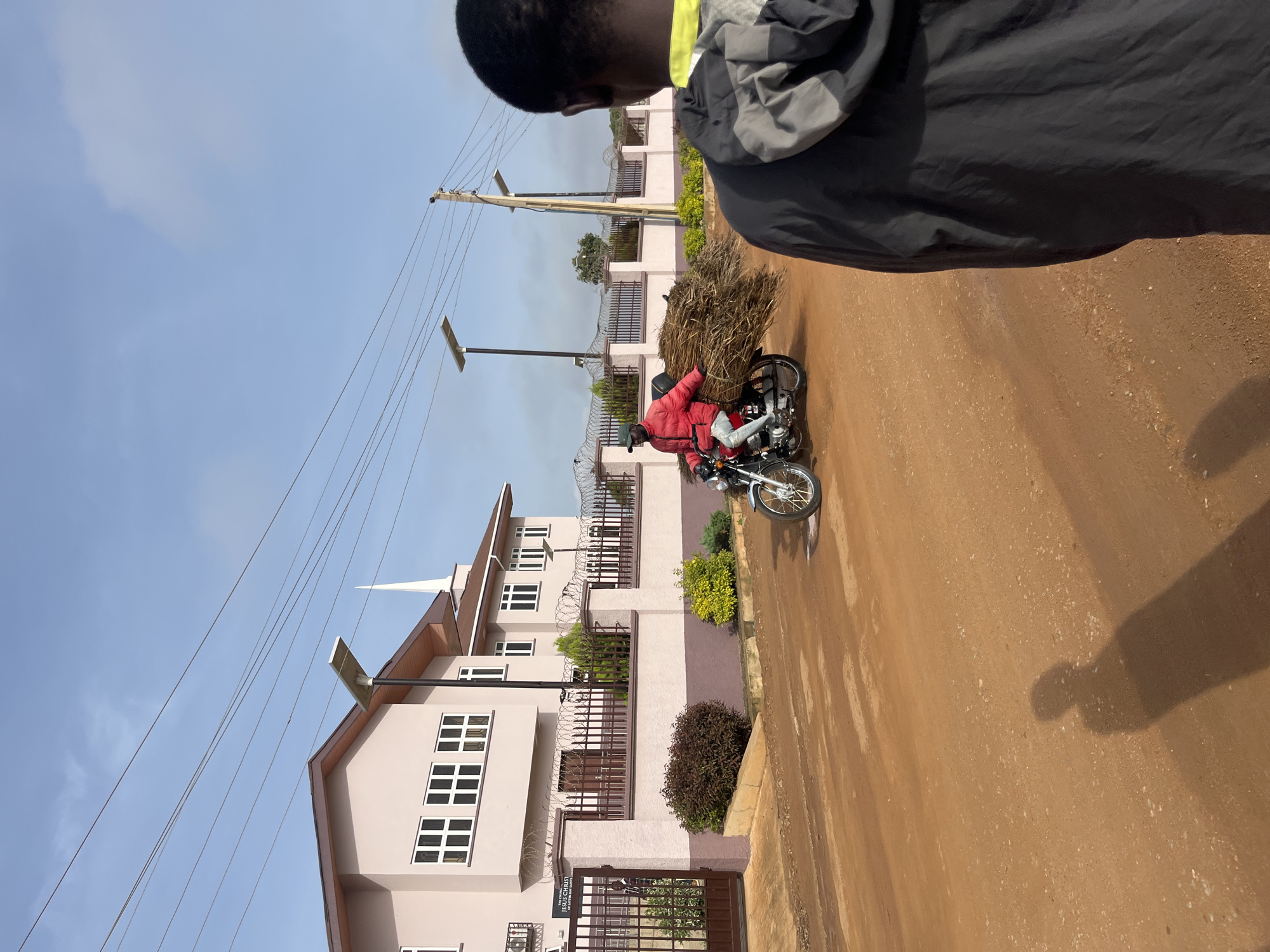 In the city of Bukuru, Nigeria, a person wearing a red jacket and a green cap rides a motorcycle laden with a large bundle of dry grass down a dirt road. The motorcycle is a light color, possibly silver, and has a visible chrome-finished wheel. To the right of the motorcycle rider, a person is seen from behind, facing away from the camera, wearing a dark jacket with a bright yellow-green collar.
Behind the motorcycle rider, a light pink building with a brown roof stands prominently. The building has multiple windows with white frames and a prominent steeple on its roof. A dark metal fence, topped with barbed wire, encloses the property. Solar-powered streetlights are visible on poles along the street. The road surface is composed of reddish-brown dirt with scattered small stones and pebbles. The sky is a pale blue with sparse white clouds. The overall scene depicts a typical street in a Nigerian city, with a blend of residential architecture, ongoing transportation, and everyday activity.