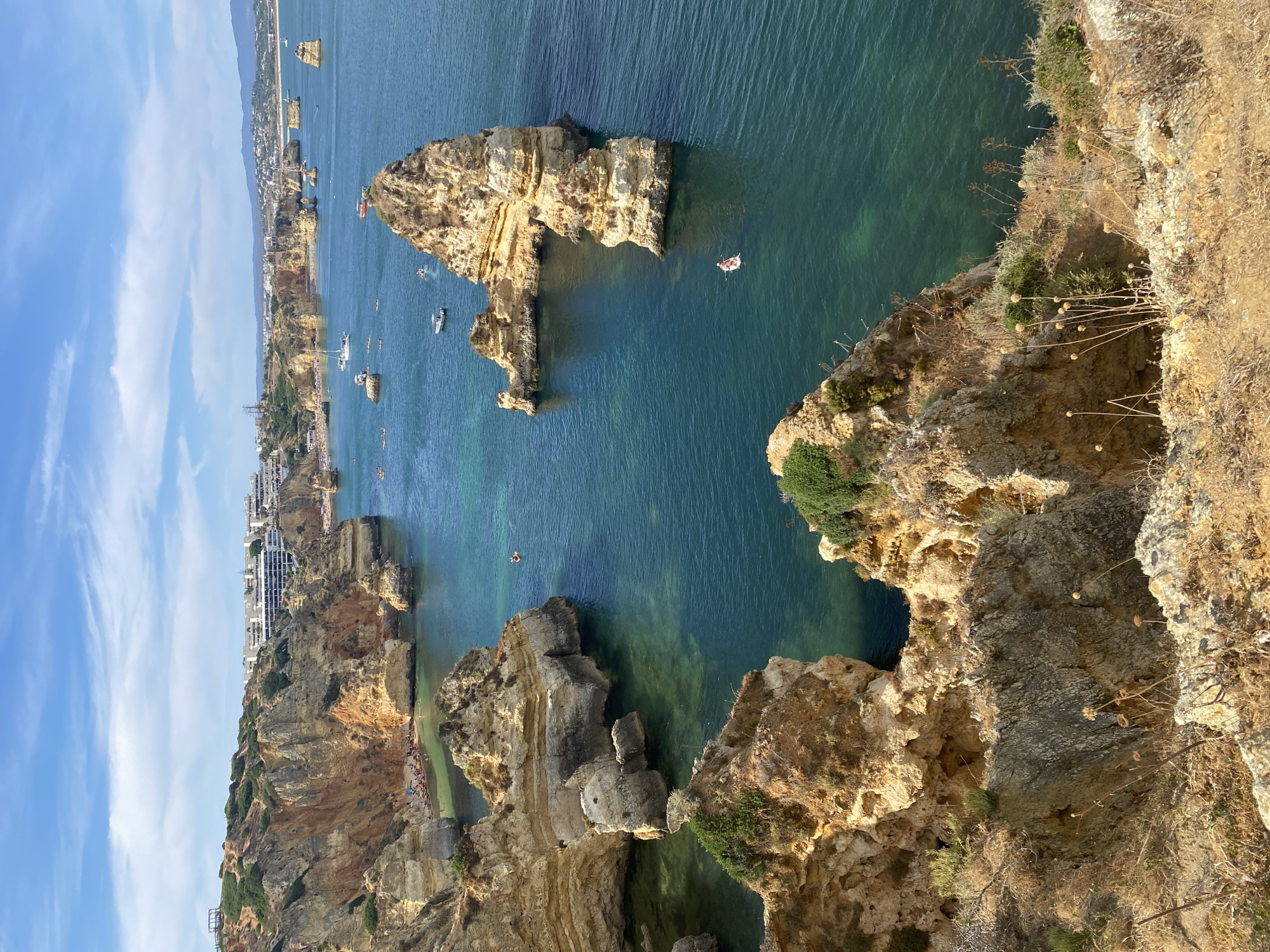 This is a high-angle view of the coastline in Lagos, Portugal, specifically the Ponta da Piedade rock formations. The image captures rugged, golden-brown limestone cliffs carved by the sea into arches, sea stacks, and grottoes. The water is a clear, vibrant turquoise, revealing sandy seabed in shallower areas. Several small boats, including a white sailboat and a few motorboats, are scattered on the water. A person is seen paddleboarding near a large rock formation in the middle of the frame. The shoreline in the background features a bustling beach with numerous sunbathers and a town with white, multi-story buildings and hotels constructed on the cliff tops. Vegetation, including dry grasses and some green shrubs, clings to the cliff faces and the foreground. The sky is bright blue with scattered white clouds.
