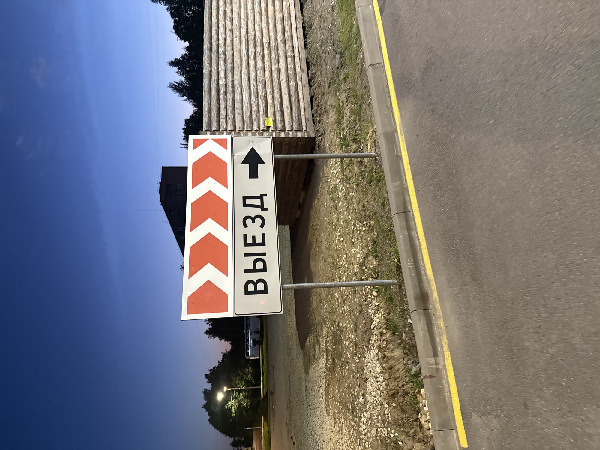 In Krasnoye Selo, Russia, a street sign with red chevron markings and the Russian word "ВЫЕЗД" (Vyyezd), meaning "Exit," is visible. A black arrow points to the right on the sign, indicating the direction of the exit. The sign is mounted on a metal pole and is positioned in front of a wooden structure constructed from logs. The scene is set against a twilight sky with a deep blue hue. In the background, trees and a building are silhouetted against the sky. The foreground shows a paved road with a yellow line marking its edge, and a curb. The area around the sign is covered with gravel and some patches of grass.