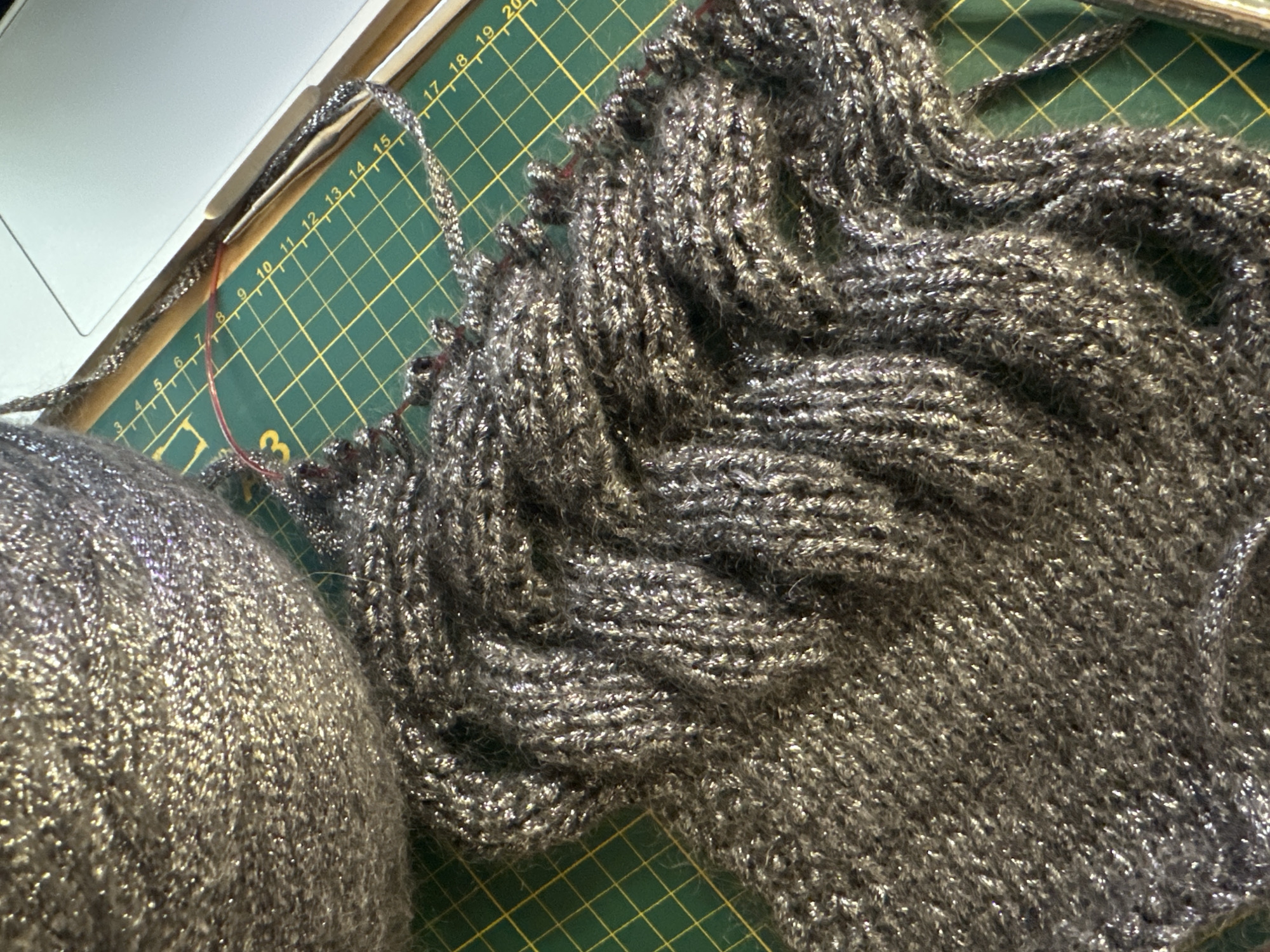 An overhead, close-up shot shows a ball of sparkly gray yarn resting on a green cutting mat with a white laptop in the background. The yarn is in the process of being knitted, with a section of the gray, sparkly fabric draped over the mat. Red knitting needles are visible, holding the knitted material. The cutting mat has a grid pattern with yellow lines and numbers indicating measurements.