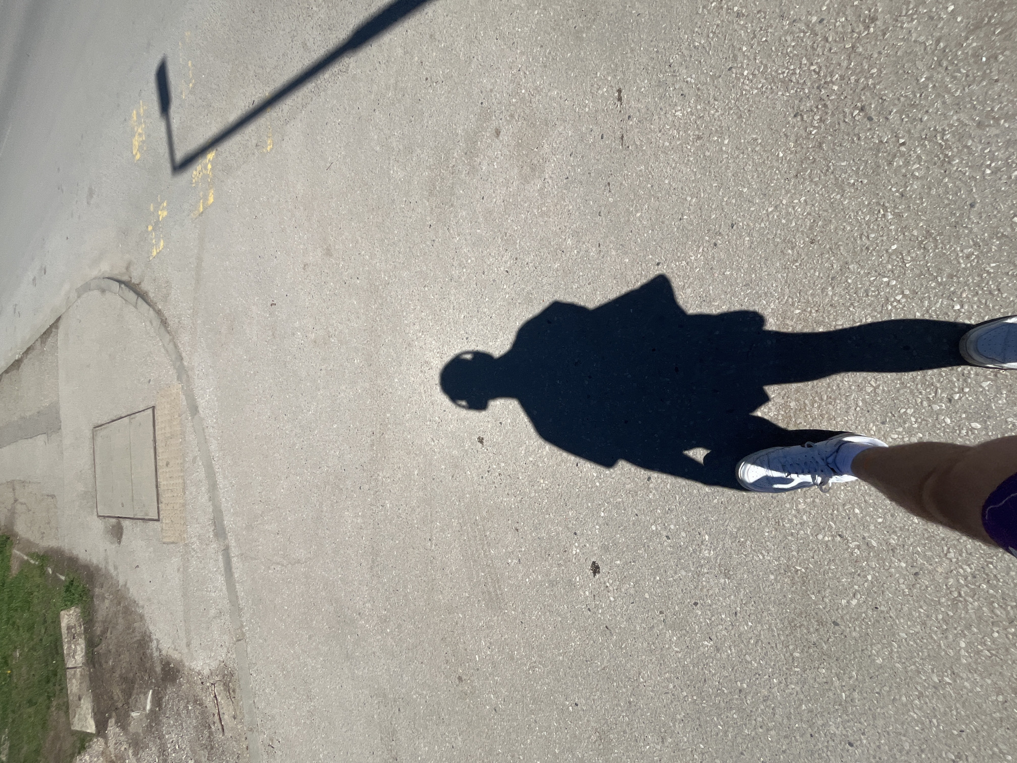 The image shows a shadow cast on a paved surface, likely a sidewalk or street, by a person wearing a helmet, t-shirt, and leggings or tight pants. The person is wearing white athletic shoes and is standing on the edge of the frame, only partially visible. The shadow of a streetlight is also visible on the paved surface. The paved surface is adjacent to a curb and a patch of grass. The location is likely Sofia, Bulgaria given the composition and the context of the original question.