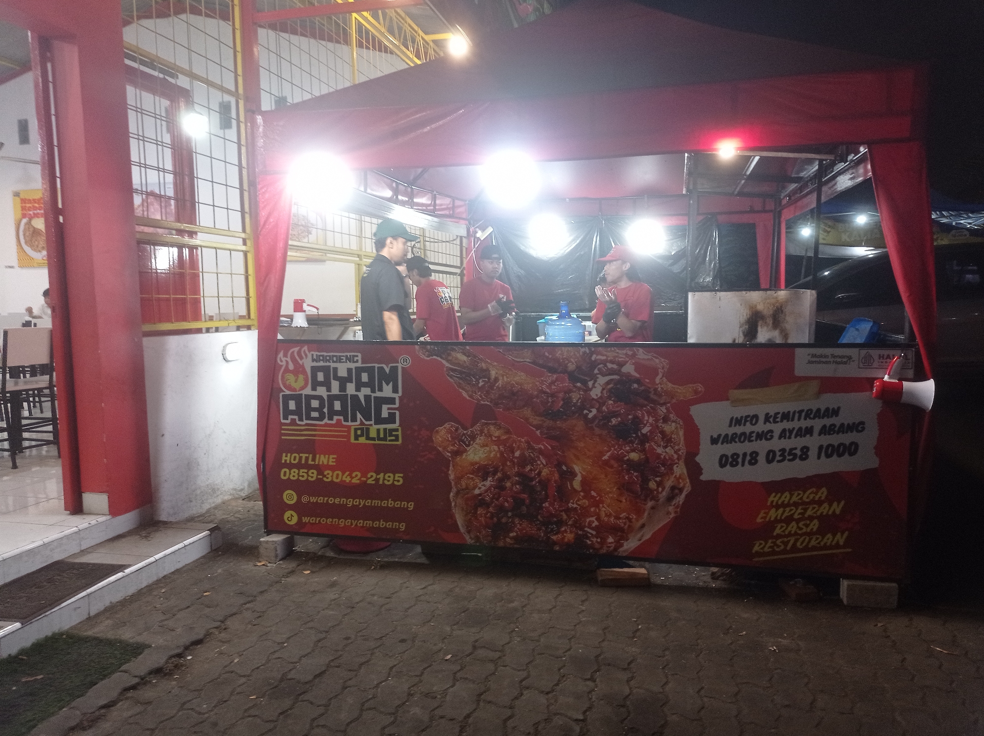 A nighttime outdoor scene at a food stall in Landungsari, Indonesia, features a "Warung Ayam Abang Plus" stall with a red canopy. Three individuals, two men and one boy, are visible behind the counter. One man in a green cap and black shirt is facing right, talking to a boy in a red shirt who is looking towards the left. Another man in a red shirt and red cap is standing in the middle, holding a microphone and looking forward, while a third man in a red shirt and red cap stands to his right, clapping. The stall's front display showcases a large image of fried chicken and promotional text in Indonesian, including contact information. Various food preparation items, such as a blue water dispenser and a metal container, are present. The ground is paved with interlocking bricks, and a small grassy area is to the left. The overall ambiance is that of a bustling local eatery at night.
