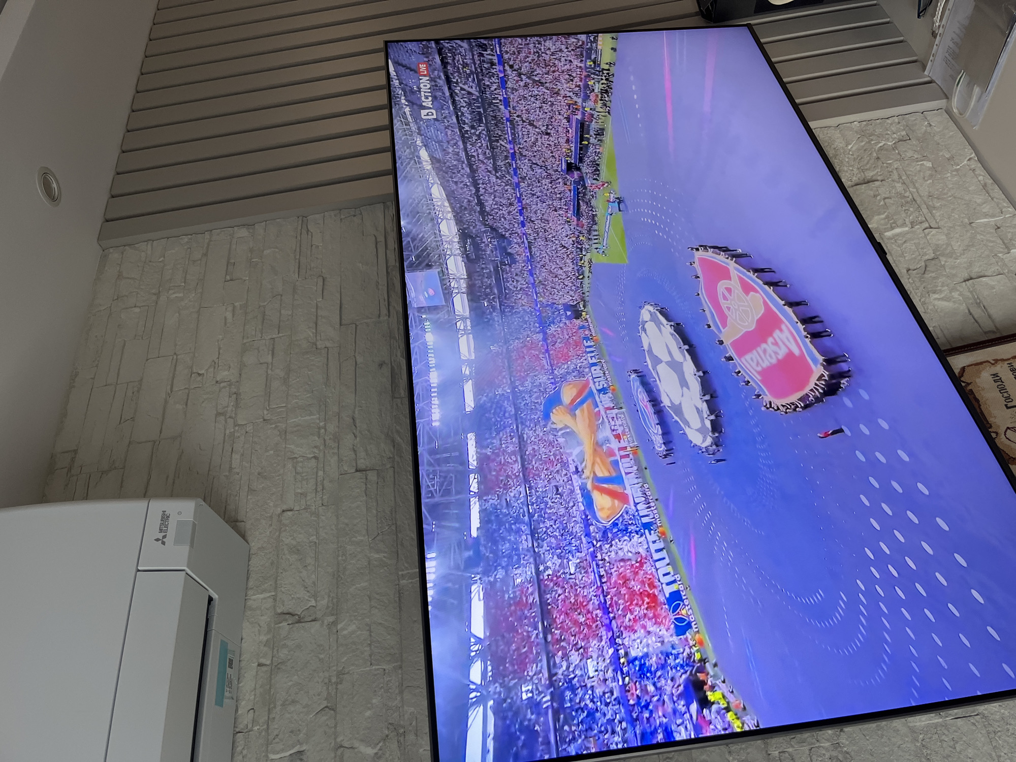 The image shows a large television screen displaying a soccer game. The screen is mounted on a wall with a textured, brick-like surface. Above the screen is a section of wall with horizontal gray slats. To the left of the screen is a white Mitsubishi Electric air conditioning unit mounted on the same wall. The image on the screen shows a soccer stadium filled with spectators, with the Arsenal logo prominently displayed. The image appears to be taken in an interior setting in Varna, Bulgaria.