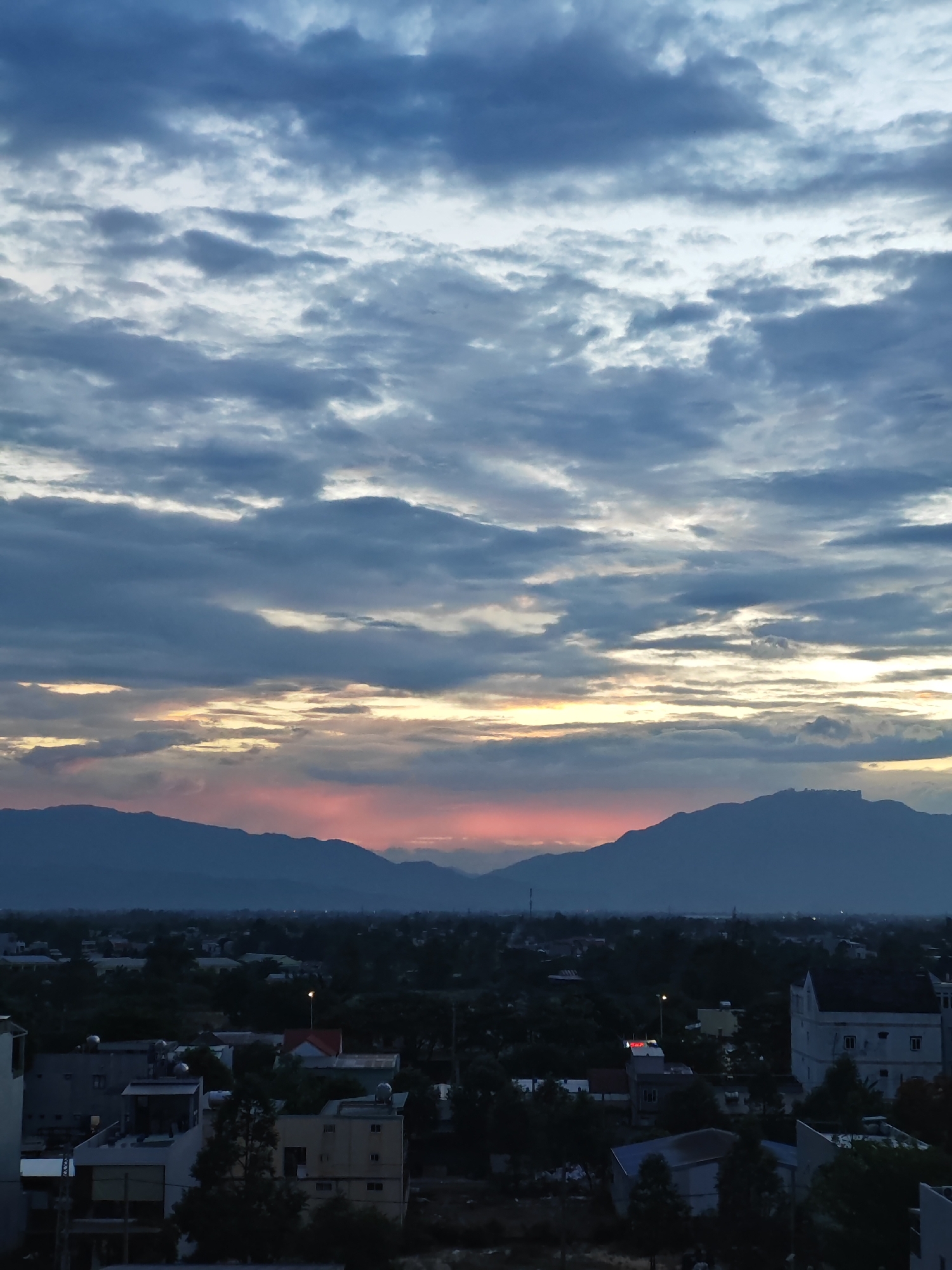 This is a vertical, eye-level, medium shot of a city skyline at dusk, likely from a high vantage point, looking towards a range of mountains. The sky is filled with dramatic clouds, ranging from deep blues and grays to lighter whites and grays, with streaks of orange and pink near the horizon where the sun is setting behind the mountains. The clouds appear textured and layered, suggesting movement and depth.
Below the sky, the mountain range stretches across the middle ground, silhouetted against the colorful sky. The mountains are a deep blue-gray, with some appearing jagged and others more rounded. A faint pink glow is visible on the lower slopes of the mountains, indicating the last rays of sunlight.
In the foreground, the cityscape is visible. Buildings of varying sizes and architectural styles are clustered together, with rooftops and upper stories clearly discernible. Some buildings are painted in light colors, while others are darker. There are also numerous trees scattered throughout the urban landscape, appearing as dark masses against the lighter buildings and sky. Streetlights are visible, some casting a yellow glow. The overall impression is one of a densely populated urban area nestled at the foot of a majestic mountain range, bathed in the soft, fading light of sunset. The scene evokes a sense of peace and natural beauty combined with human habitation.