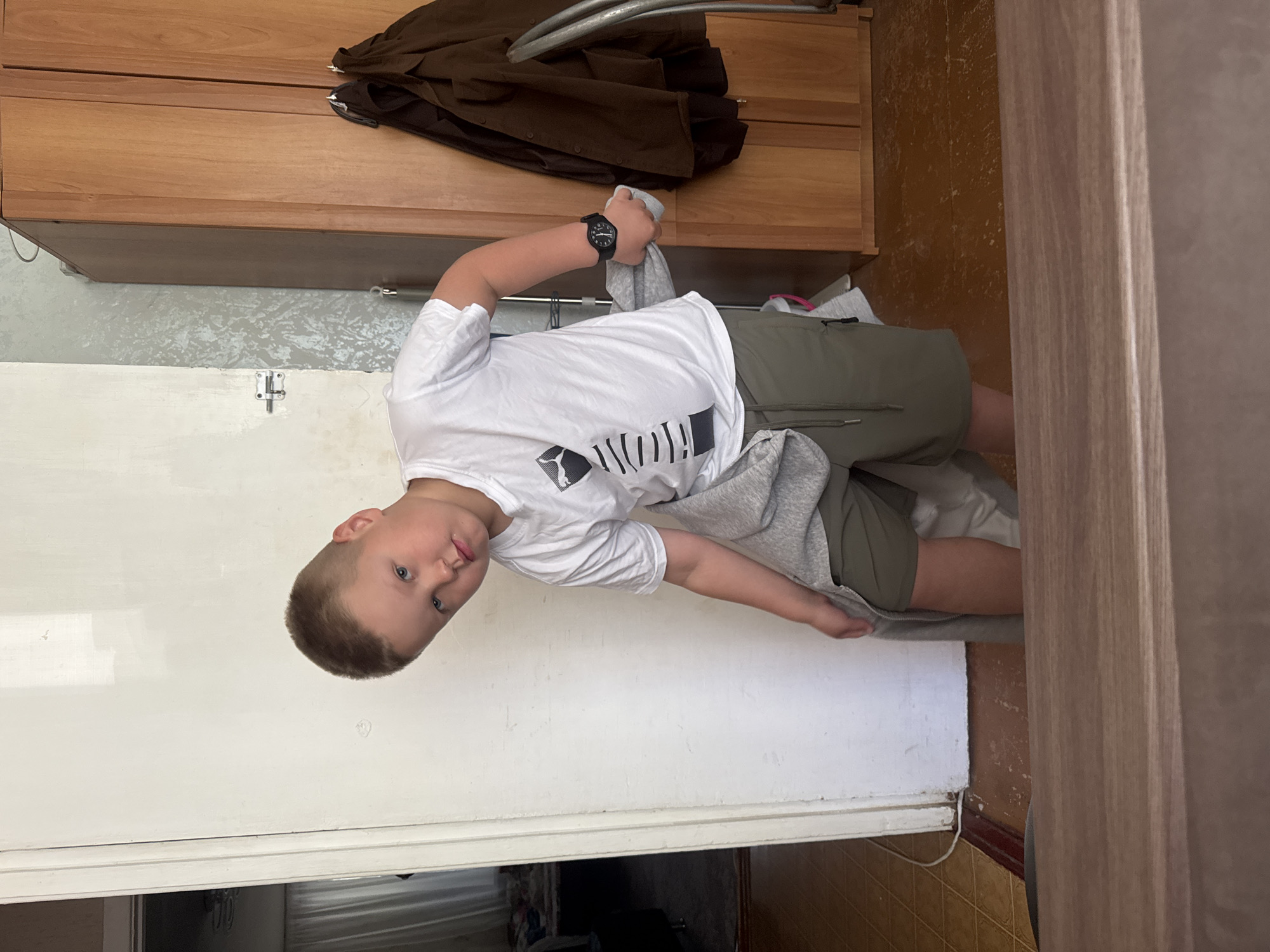 A young boy with short brown hair and blue eyes is standing in what appears to be a room. He is wearing a white t-shirt with a black and white graphic design and olive green shorts. The boy is holding a grey piece of clothing in his left hand and appears to be pulling it down. He is looking towards the camera with his head tilted.
In the background, there is a white door on the left side of the image, partially ajar, revealing a glimpse of another room with a curtain and some furniture. To the right of the door, the wall has a textured grey wallpaper. Further to the right, there is a tall wooden wardrobe with two doors. Two dark-colored jackets are hanging on the doors of the wardrobe. The floor is made of linoleum tiles. A wooden surface, possibly a table or counter, is visible in the foreground.
Based on the provided context of the city being Chaikovsky, Russia, this scene depicts an indoor setting, likely a residential space within the city. The overall impression is casual and domestic.