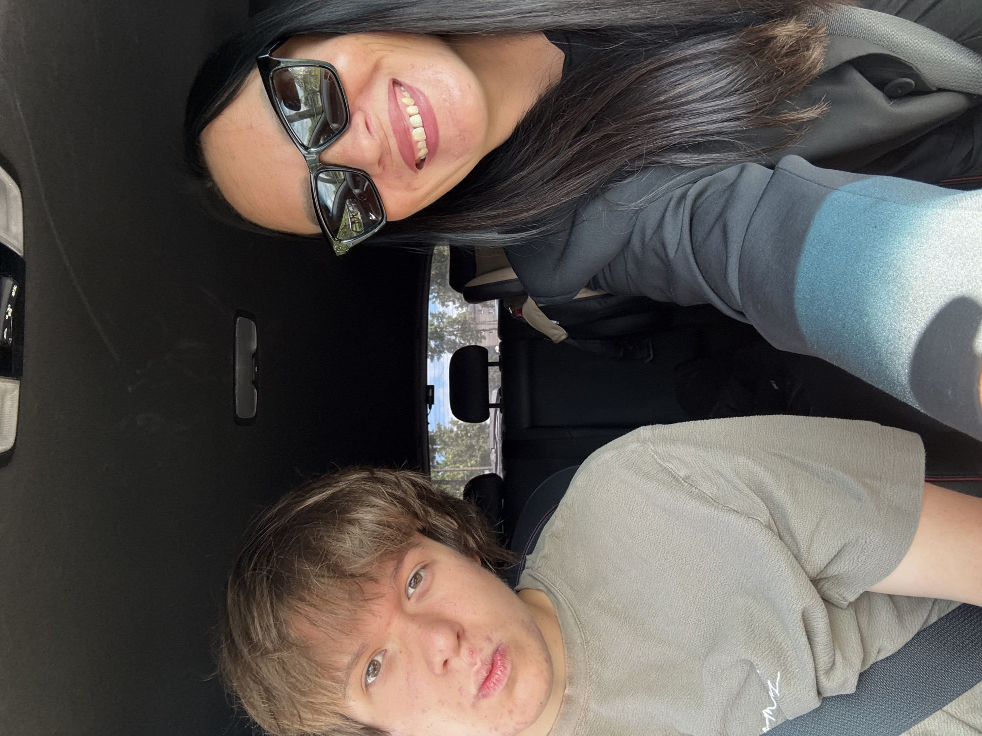 Two people are inside a vehicle. On the left is a young person with light brown hair, wearing a grey t-shirt and a seatbelt. Their facial expression is neutral, with a slight pout. On the right is an adult with long, dark hair, wearing sunglasses and a black jacket. They are smiling broadly. The interior of the car, including black seats and the ceiling, is visible. The background outside the car shows trees and buildings, suggesting an outdoor environment, possibly within a city, although its specific location in Chaikovsky, Russia, cannot be determined from the image alone.
