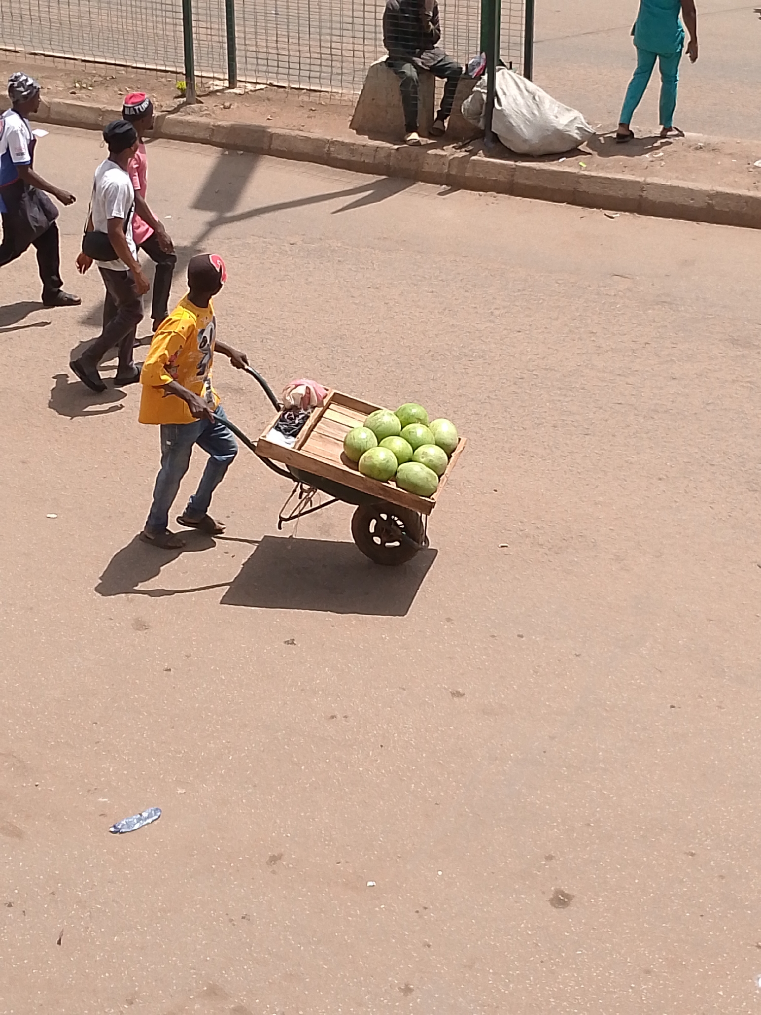 A street in Bukuru, Nigeria, shows a young man pulling a wooden cart laden with approximately a dozen green watermelons. He wears a yellow shirt, blue jeans, and a red head covering. The cart has a single wheel. Several other individuals walk on the sidewalk in the background. A piece of litter lies on the road surface.