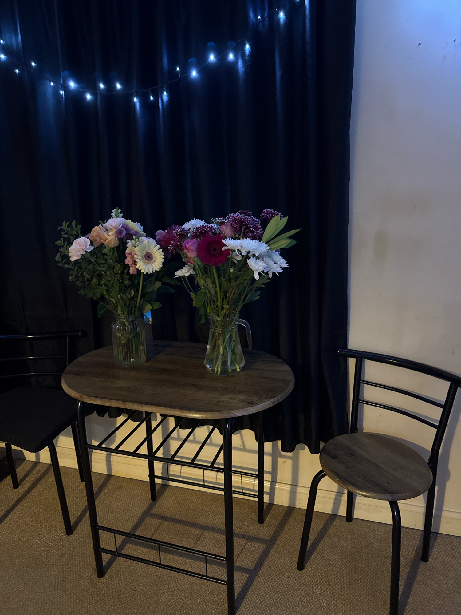The image depicts a small, intimate dining setup with two chairs flanking a round wooden table. Two glass vases filled with colorful flower arrangements are placed on the table. A dark curtain forms the backdrop, with a string of white fairy lights draped above it, casting a subtle glow. The overall atmosphere suggests a cozy, perhaps romantic, setting. The location is not specified in the image, and therefore it cannot be definitively placed in Great Yarmouth, United Kingdom.