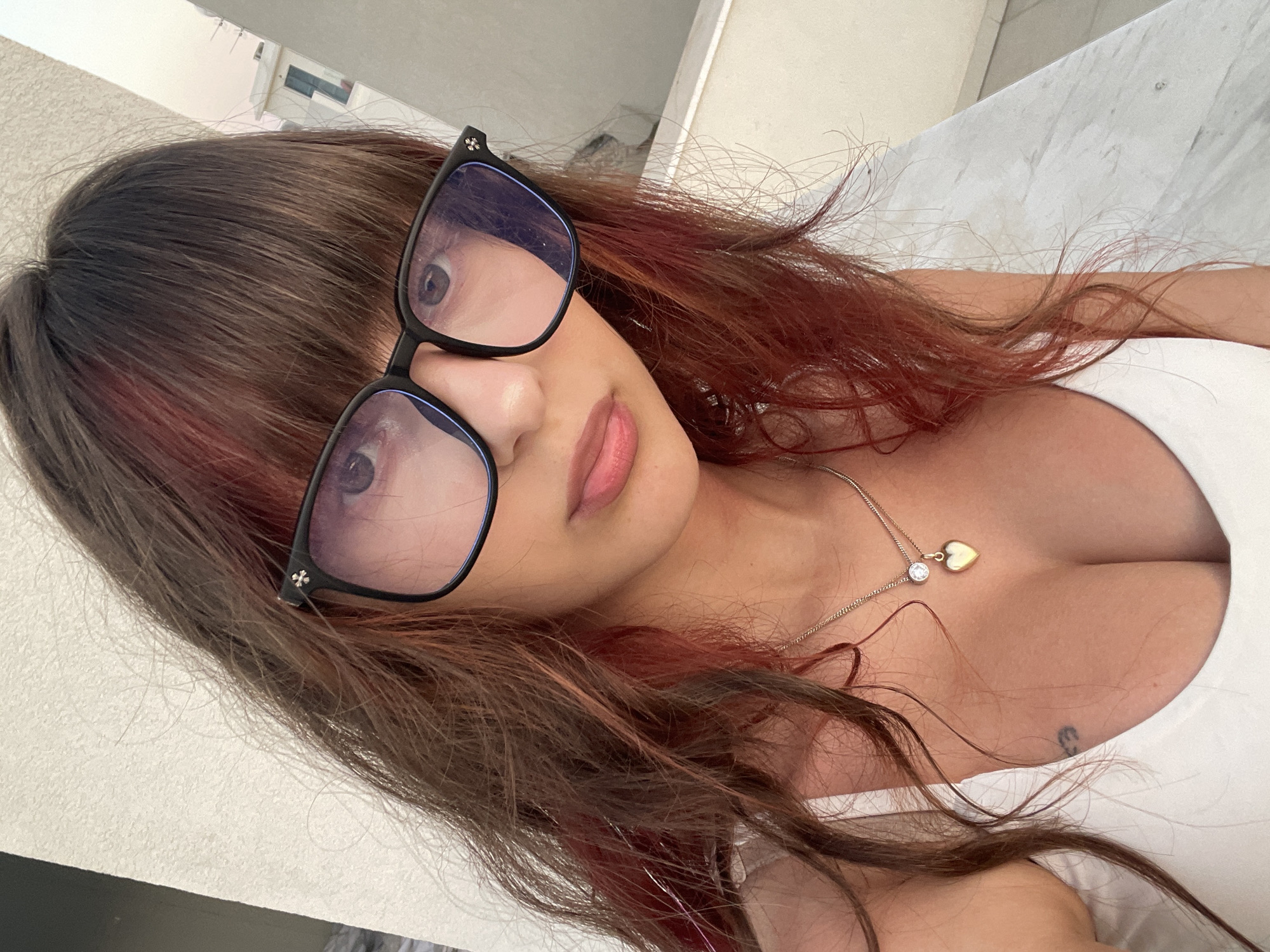 A close-up, waist-up portrait captures a young woman wearing large, black-framed glasses and a white tank top. Her long, brown hair, with streaks of red, frames her face. A delicate gold necklace with a heart-shaped pendant is visible on her chest, and a small tattoo is located on her left side. The background consists of a textured, light-colored wall and hints of buildings and the sky in the distance, suggesting an outdoor urban setting, possibly a balcony or rooftop in Paphos Municipality, Cyprus. The lighting indicates it may be late afternoon or early evening.