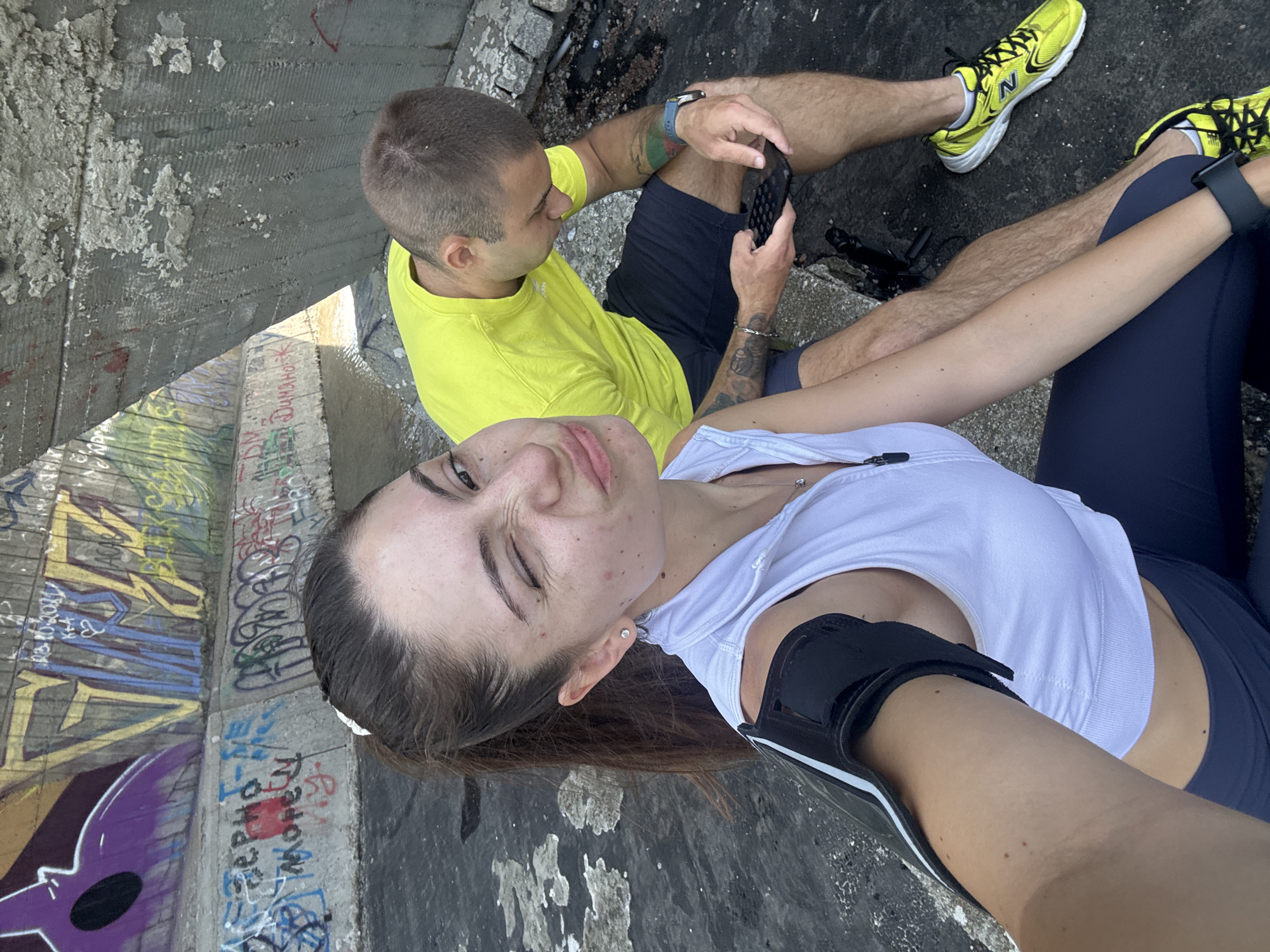 A man and a woman, dressed in athletic wear, are sitting outdoors in front of graffiti-covered concrete structures. The woman is in the foreground, taking a selfie and winking at the camera. She has her hair in a ponytail and is wearing a white zip-up vest over a white sports bra and dark leggings. An armband is visible on her left arm. The man is in the background, looking down at his phone, and is wearing a bright yellow t-shirt and dark shorts. He has tattoos on his arms and is wearing a watch. The background consists of a concrete embankment with various graffiti tags and drawings, and a body of water. This scene appears to be located in Varna, Bulgaria, likely near the Varna beach promenade or a similar urban coastal area with graffiti art.