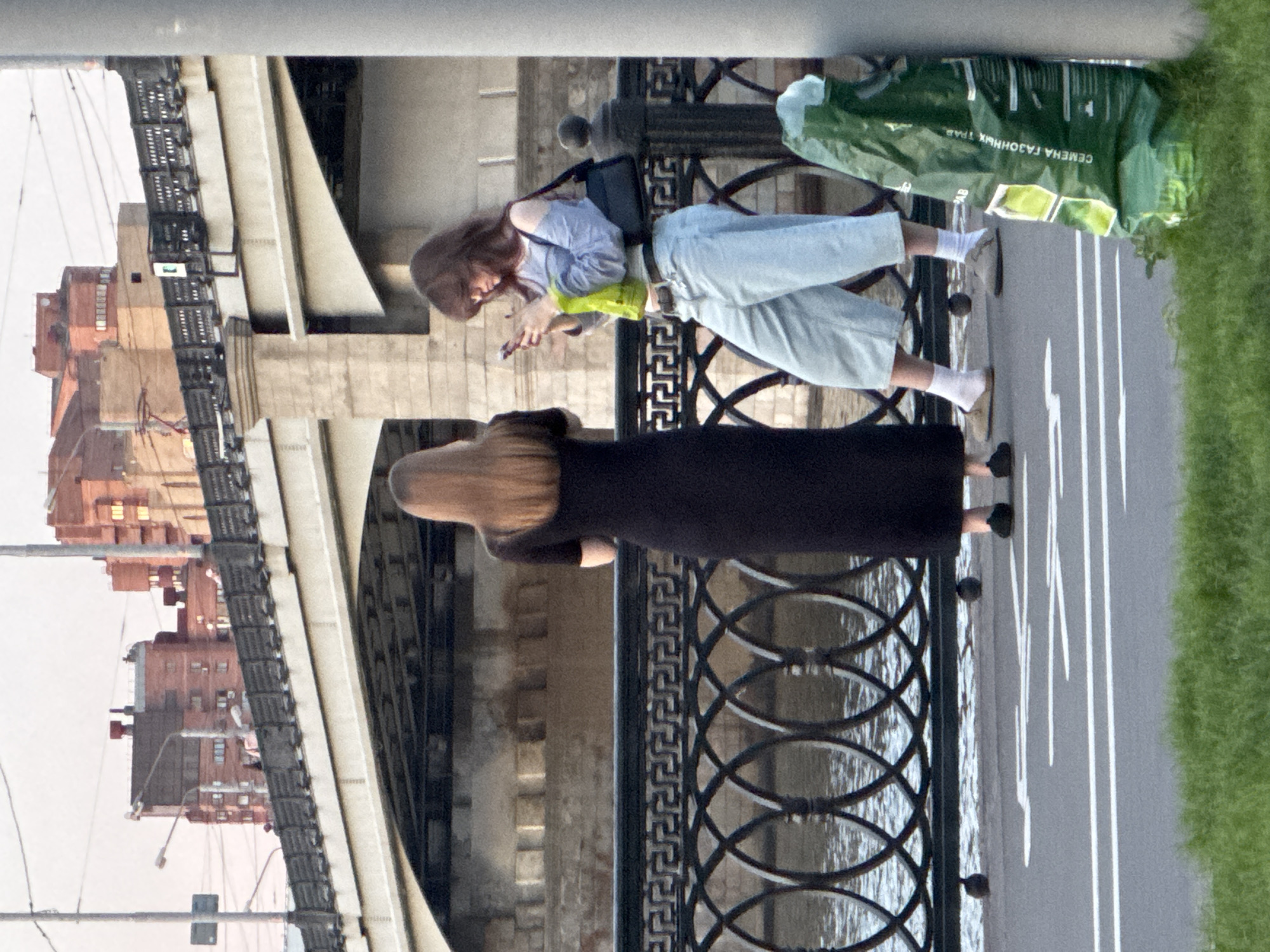 The image captures an urban waterfront scene in Unknown, Russia, featuring two women on an embankment walkway beside a river or canal, with a large bridge and buildings in the background.
To the left, a woman with long, straight, light brown hair, seen from behind, stands next to a decorative black metal railing. She is wearing a form-fitting, short-sleeved, floor-length dark dress and dark flat shoes, looking towards the water.
To her right, a second woman with medium-length, wavy dark brown hair is seen in a three-quarter profile, looking down at a smartphone held in both hands. She wears a light blue, off-the-shoulder long-sleeved top, light wash baggy jeans, white socks, and light-colored slip-on shoes. A black shoulder bag hangs from her left shoulder, and she holds a bright green plastic bag in her left hand.
The black metal railing, featuring repeating circular and geometric patterns, separates the paved walkway from the water, which shows ripples on its surface. White lines and partial symbols, indicative of a bicycle lane, are painted on the grey paved surface in the foreground. Green grass lines the very bottom edge of the frame. In the lower right foreground, a large, crumpled green plastic bag with Russian text "СЕМЕНА ГАЗОННЫХ ТРАВ" (LAWN GRASS SEEDS) printed on it leans against a grey pole.
In the mid-ground, a multi-arched bridge with stone piers and a black metal railing spans the background. Beyond the bridge, several multi-story buildings, predominantly reddish-brown brick, are visible under a light, hazy sky. Some windows in the background buildings appear to be illuminated. The scene is illuminated by soft, natural light, suggesting late afternoon or early evening.