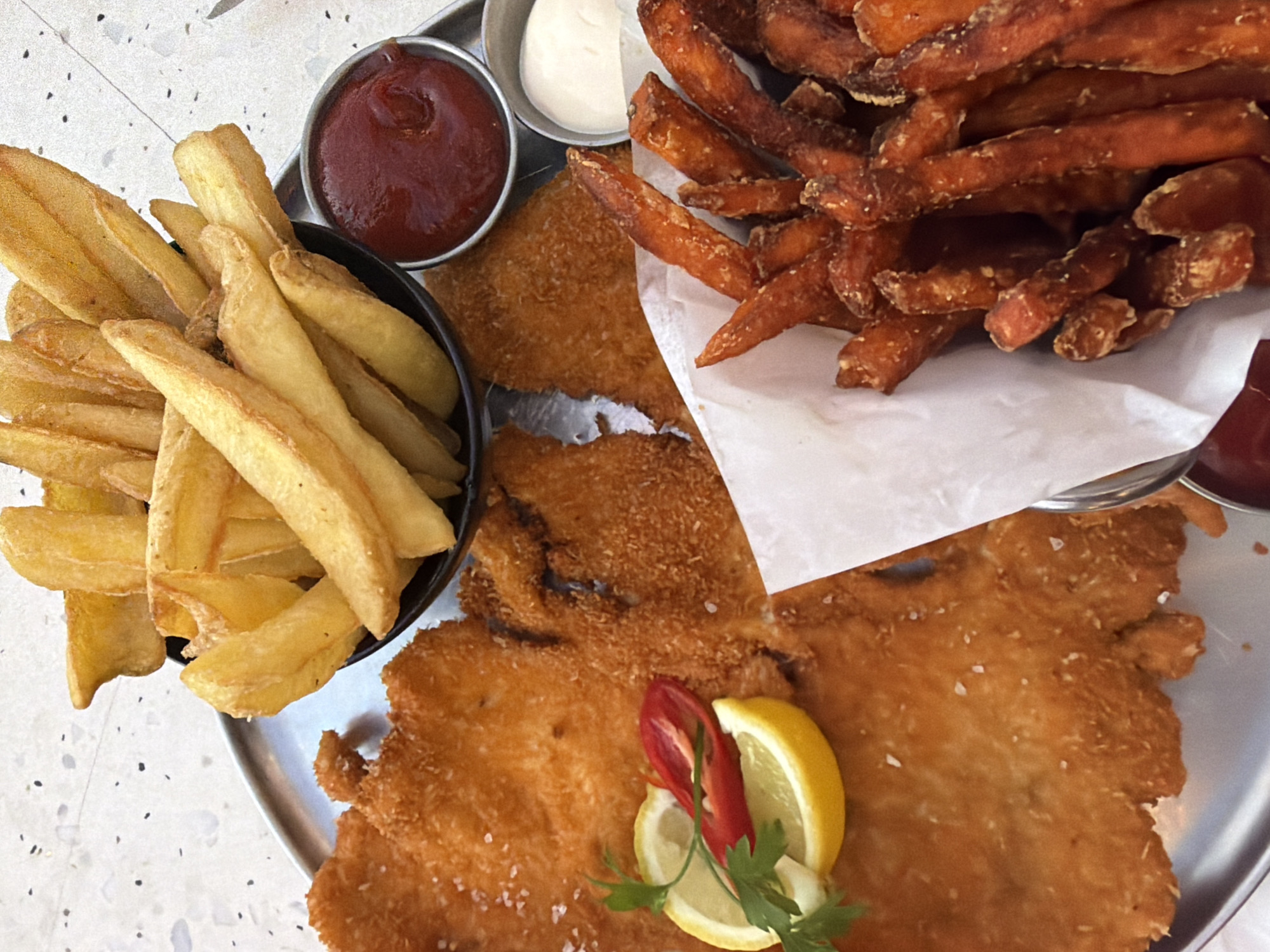 The image shows a meal served on a metal tray, likely in a restaurant in Tel-Aviv, Israel. The meal includes a schnitzel garnished with a lemon wedge, parsley, and a slice of red chili. Side dishes are regular fries, served in a small black bowl, and sweet potato fries on a piece of white paper. Two small metal ramekins containing ketchup and mayonnaise are also part of the presentation. The food is set against a background with a speckled pattern.