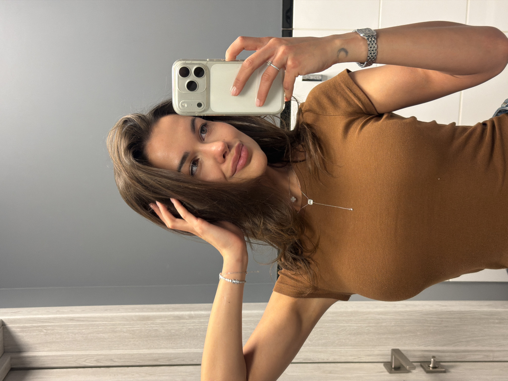 A mirror selfie features a young woman, with the image rotated 90 degrees counter-clockwise. She is looking towards the viewer with a slight smile and full lips. Her brown hair falls around her shoulders, and her left hand is gently placed in her hair near her head, while her right hand holds a smartphone up to capture the reflection.
The smartphone appears to be an iPhone, visible through a clear protective case that reveals its triple-lens camera system on the back. On her right hand, a silver ring adorns her ring finger, and a classic silver-toned watch is worn on her right wrist. A small crescent moon tattoo is visible on the inner side of her right forearm. Her left wrist displays a delicate silver bracelet. She is wearing a fitted, ribbed, short-sleeved brown t-shirt and a subtle silver necklace with a small square pendant.
The background, reflected in the mirror, shows a minimalist setting. A plain gray wall forms the upper portion, meeting a light-colored, wood-textured counter or shelf in the lower part of the frame. In the bottom right corner of the reflected counter, metallic fixtures are visible, possibly indicating a bathroom or similar utility space. The overall impression is one of a clean, modern indoor environment in Krasnoyarsk, Russia. There is no visible text in the image.