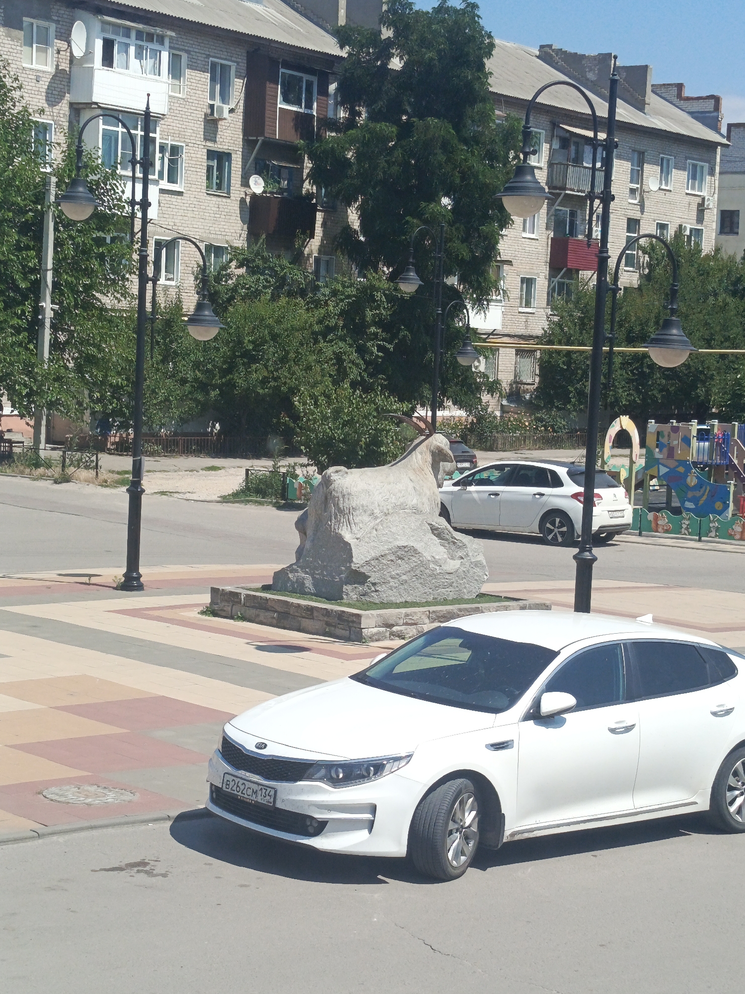 The image shows a street scene in Uryupinsk, Russia, on a sunny day. In the foreground, a white Kia Optima sedan is parked on the right side of the road, facing towards the viewer. The car has a license plate that reads "B262CM 134". Behind the Kia, a white hatchback is parked in the street. A large stone sculpture of a goat stands on the left side of the road, near a sidewalk paved with colorful tiles. In the background, a multi-story apartment building with light-colored brick walls is visible. Trees and streetlights line the street. To the right of the apartment building, a colorful children's playground with slides and other equipment is present.