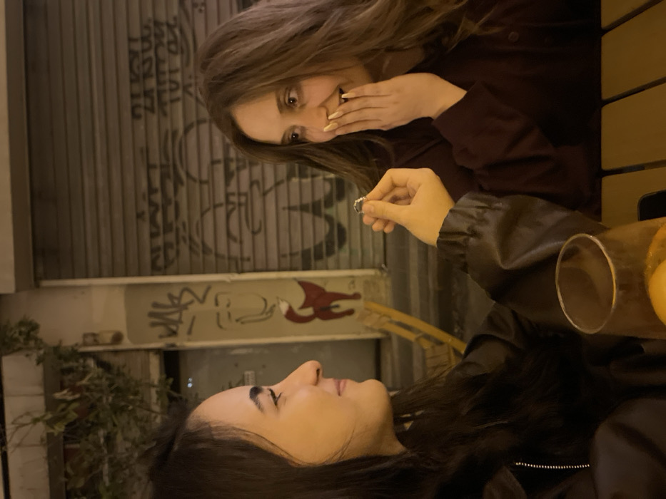 Two women are seated at an outdoor table. One woman, facing forward, is holding up a ring. The other woman, facing her and slightly behind, reacts with surprise, her hand to her mouth. Behind them is a building with graffiti and a closed rolling gate. A drink sits on the table. The scene appears to be a proposal or a similar event.