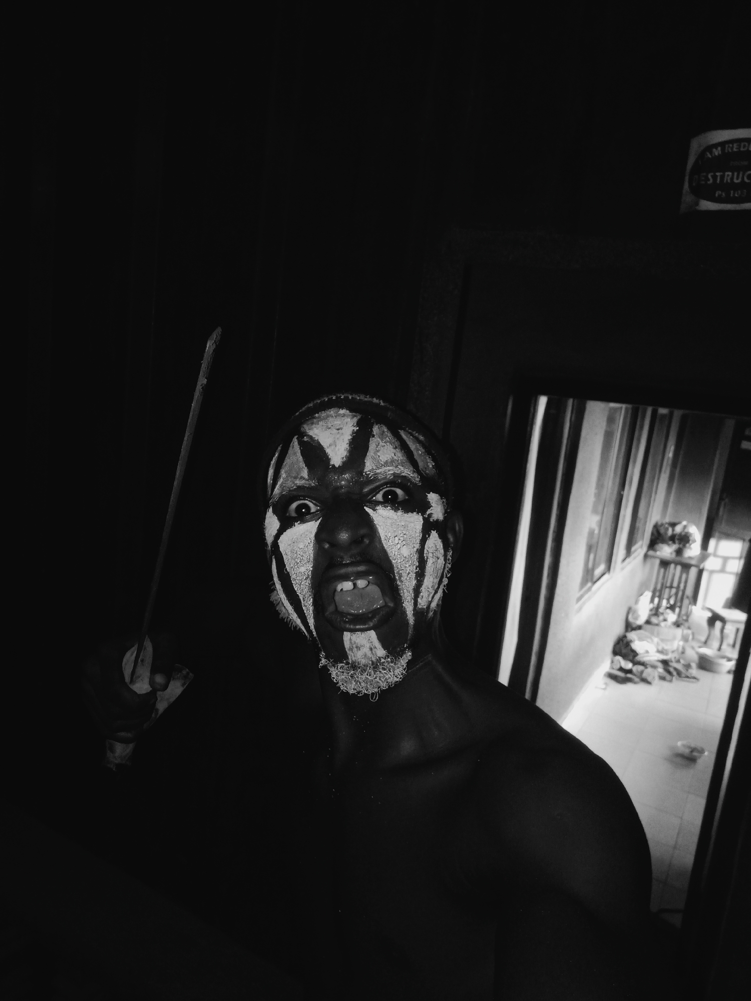 This striking black and white photograph, likely taken in Onitsha, Nigeria, features a person with elaborate face paint, captured in a moment of intense expression. The subject, appearing male, is positioned in a dark, interior space, looking directly into the camera. Their face is adorned with bold white and black patterns, resembling tribal or ceremonial markings, which strongly contrast with their dark skin.
The individual's eyes are wide and piercing, and their mouth is open in what appears to be a scream, yell, or dramatic vocalization, revealing their teeth. In their left hand, they hold a long, slender, stick-like object, possibly a rod or a symbolic weapon, held upright. Their upper body is bare, further emphasizing the dramatic face paint and intense expression.
To the right of the subject, a doorway or opening reveals a brighter background, showing an interior room or a covered outdoor area. This space appears somewhat cluttered with visible furniture, windows, and various items on the floor, suggesting a domestic or semi-public setting.
In the upper right corner, a partially visible sign or label displays fragmented text, including "AM REDU", "DESTRUC", and "Ps 103". This could be part of a warning, instruction, or a product label, potentially related to fire safety or a specific code. The overall mood is powerful and theatrical, hinting at a performance, ritual, or a moment of raw emotion.