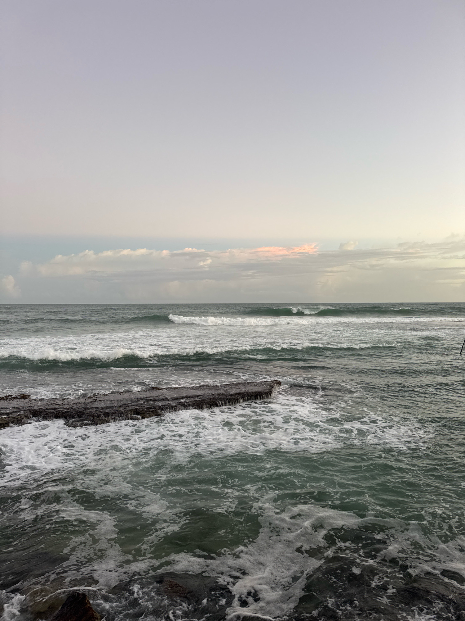 The image depicts a tranquil coastal scene in Goviyapana, Sri Lanka, at dusk. The sky above is a soft gradient of pale lavender and grey, with a hint of pale pink in the distance. Wispy clouds dot the horizon. Below, the ocean is a deep, murky green, with gentle waves lapping against a rocky shore in the foreground. White foam from the waves washes over the dark, textured rocks. The water stretches out to meet the sky, with the horizon line clearly defined. No people or specific objects are visible apart from the natural elements of the sea and sky.