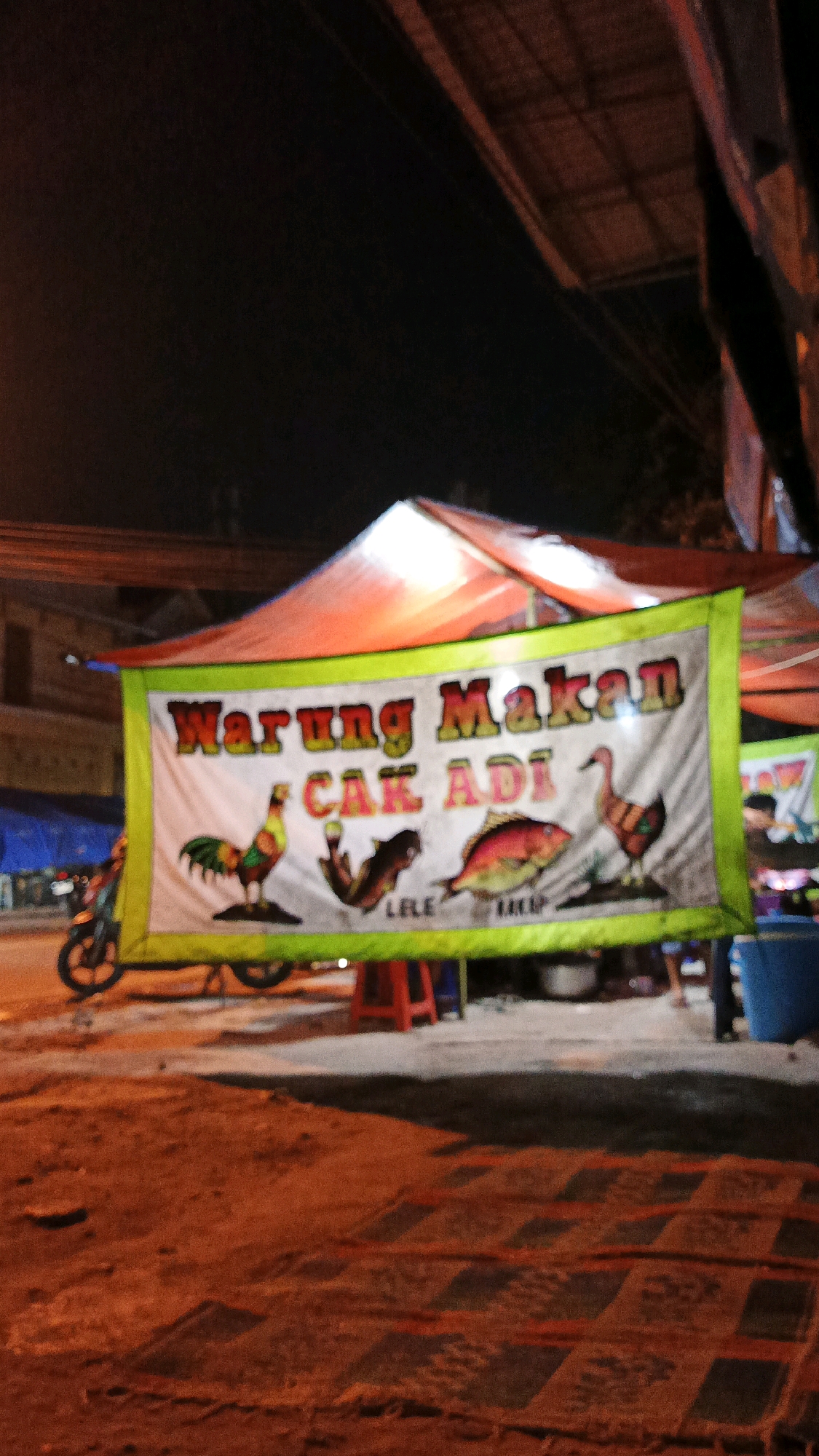 An outdoor food stall, identified as "Warung Makan Cak Adi," is visible at night in Surakarta, Indonesia. A large banner with green trim displays the stall's name in red and yellow lettering. The banner also features illustrations of a rooster, a catfish labeled "LELE," a fish labeled "KAKAP," and a duck. The stall is illuminated by artificial lighting, casting a warm glow on the surrounding area. In the background, a motorcycle is parked to the left of the stall, and a blue plastic container is visible to the right. Several people are present near the stall, though their faces are not clearly discernible. The ground appears to be dirt and gravel, with some areas covered by a patterned mat. The overall atmosphere suggests a bustling local eatery operating after dark.