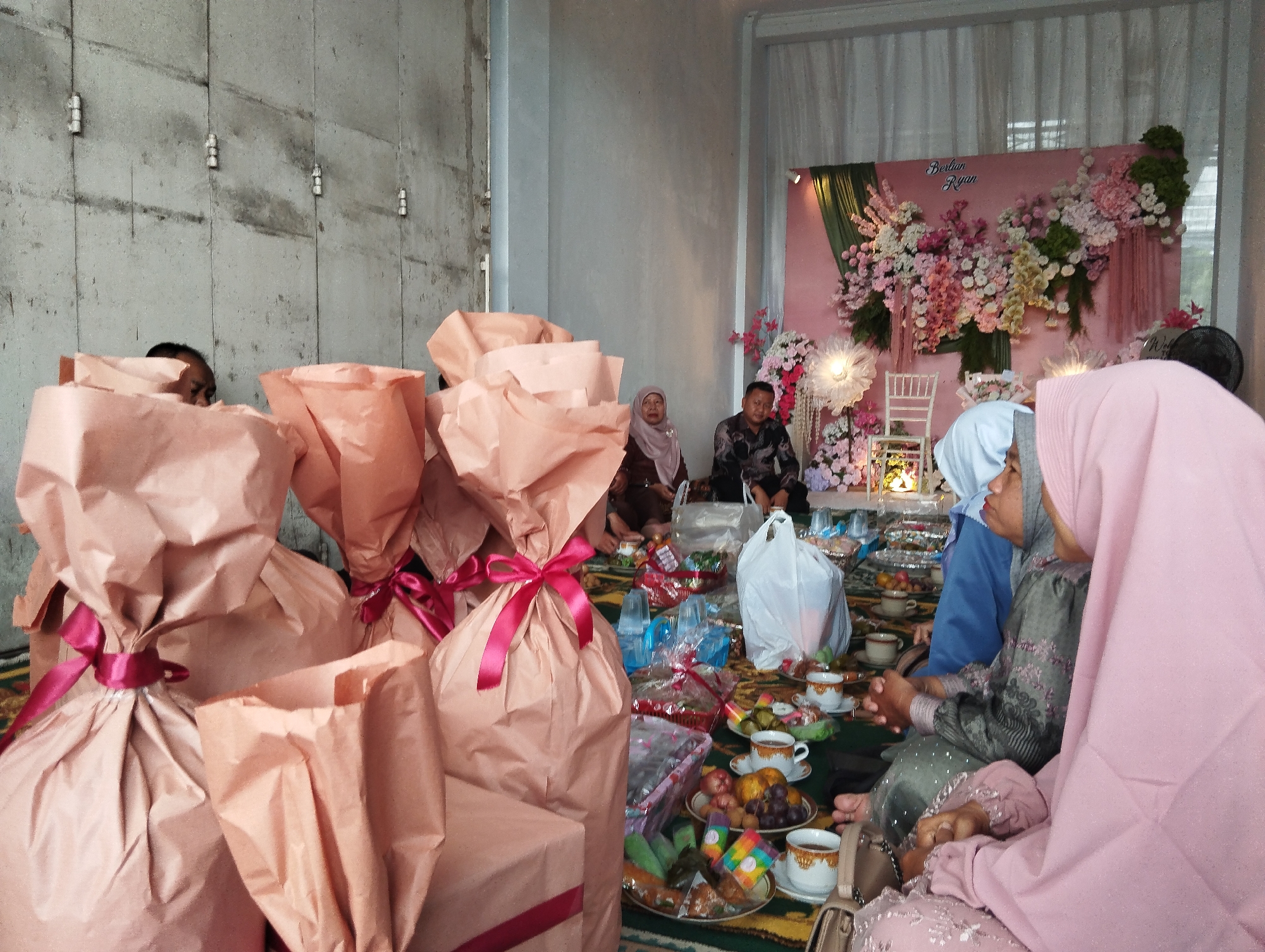 The image depicts an indoor gathering in Pangkah, Indonesia, likely a wedding or engagement ceremony, based on the floral backdrop with the names "Berlian" and "Ryan." In the foreground, several large gifts wrapped in light brown paper and tied with dark pink ribbons are stacked on the floor. A group of people are seated on the floor around a low table laden with food and drinks, including plates of fruit, pastries, and cups of tea. The people are dressed in traditional Islamic attire, with women wearing hijabs. One woman in the foreground is wearing a pink hijab and a pink patterned dress, and another is in a gray hijab and a gray embroidered dress. In the background, a man and woman are seated near the floral backdrop. The environment appears to be a decorated room with concrete walls on the left side of the frame and a curtained window on the right. The lighting suggests an indoor setting, possibly during the daytime.