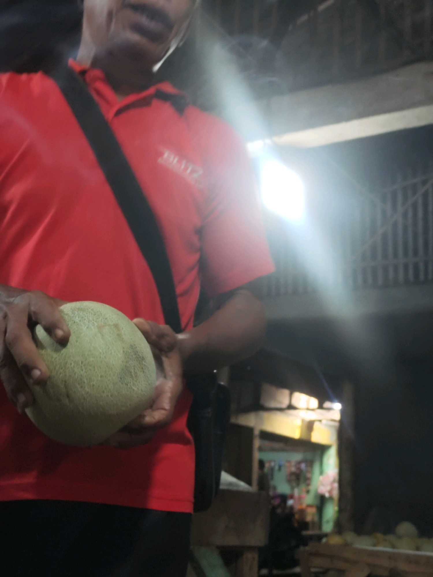A person, wearing a red shirt with a black strap across their chest and black pants, holds a light green cantaloupe in their hands. The person's face is not fully visible, but their mouth is slightly open. The background is dark and appears to be an indoor market or shop in Karawang, Indonesia. The market stalls in the background are dimly lit and show various goods, including what appear to be more cantaloupes on a wooden pallet. Bright light sources in the background create a lens flare effect.