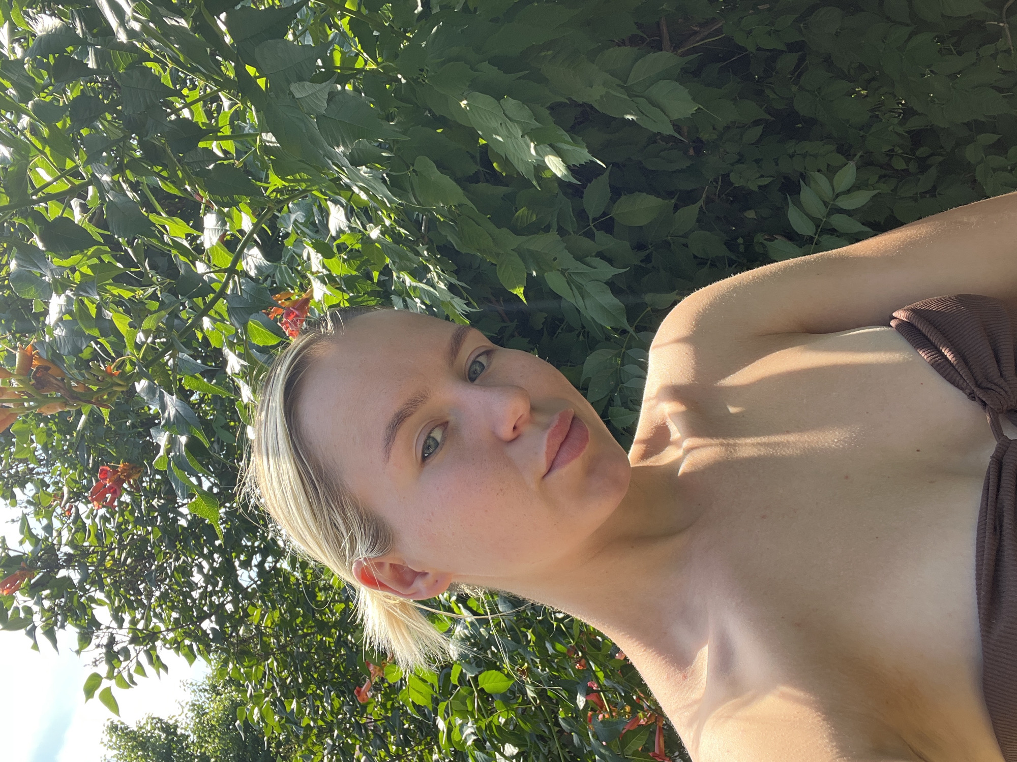 A woman with blonde hair pulled back and wearing a brown top is shown from the chest up. She is outdoors, surrounded by lush green foliage and some orange flowers. The sun is shining brightly, casting highlights on her skin and the leaves. Her head is tilted back, and she is looking towards the camera with a gentle expression. The image appears to be taken in the daytime, likely on a warm, sunny day, given the lighting and greenery. The overall mood is serene and natural. There is no visible text in the image.