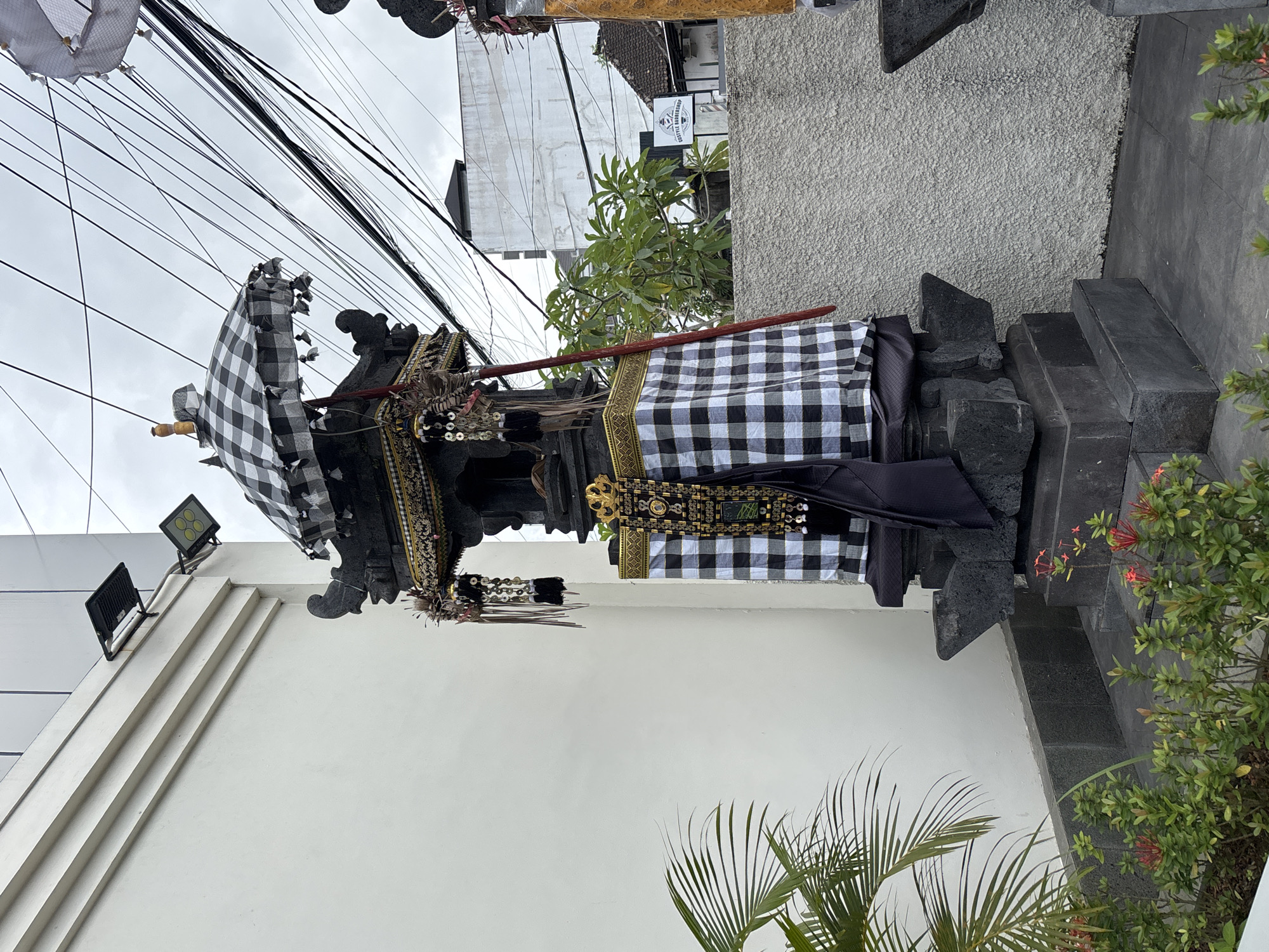 The image shows an outdoor scene in Kuta Selatan, Indonesia, featuring a decorative stone structure. The structure is elaborately adorned with a black and white checkered fabric, a gold embroidered banner, and hanging ornaments. A black and white checkered umbrella, also decorated with tassels, is positioned at the top of the structure. In the background, a white building with external lights is visible, as are tangled electrical wires stretching across the sky. A lush green plant with broad leaves is in the foreground to the left, and a garden bed with small red flowers is at the base of the structure. The overall impression is of a cultural or religious decoration in an urban or semi-urban environment.