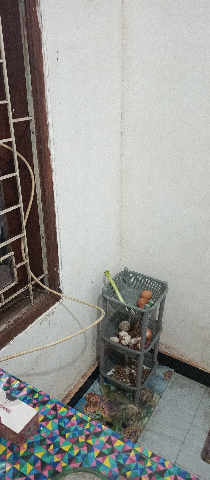 In a corner of a room, a grey plastic tiered shelf holds various items. The top shelf contains a single stalk of green celery and three brown eggs. The middle shelf holds several bulbs of garlic and one red onion. The bottom shelf appears to contain dried spices or small roots. The shelf unit is positioned on a floor covered with a grey tiled linoleum depicting a cartoon image of three girls. To the left of the shelf is a window with a dark brown wooden frame and a white metal grate. A pale yellow electrical cord snakes from the window area and curves towards the right side of the image. In the foreground, a portion of a table is visible, covered with a colorful geometric patterned cloth featuring triangles in shades of pink, blue, green, yellow, and black. A small white box with red lettering, partially obscured, rests on the table. The walls are a plain, off-white color, with a faint vertical seam visible on the right wall. The scene is indoors, likely in a domestic setting in Arjasa, Indonesia.