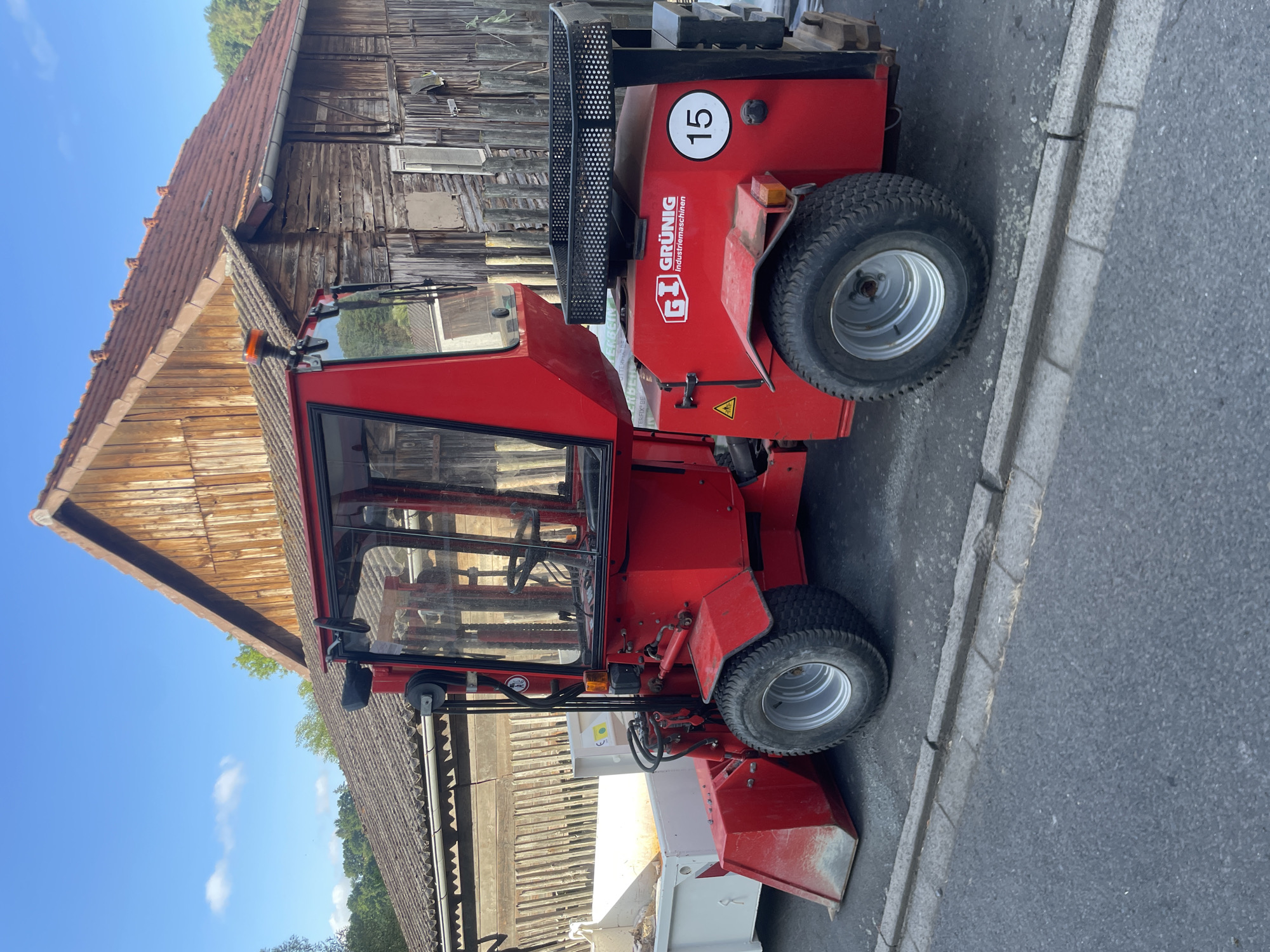 A red GRÜNIG Industriemaschinen vehicle with a white trailer is parked on a paved street in front of a rustic wooden building with a tiled roof. The vehicle has a closed cabin, large tires, and a front loader. A black basket is attached to the back of the vehicle, and the number "15" is visible on its side. The environment suggests a rural or semi-rural setting in Neukirchen, Germany, with trees and a clear blue sky in the background. No people are present in the image.
