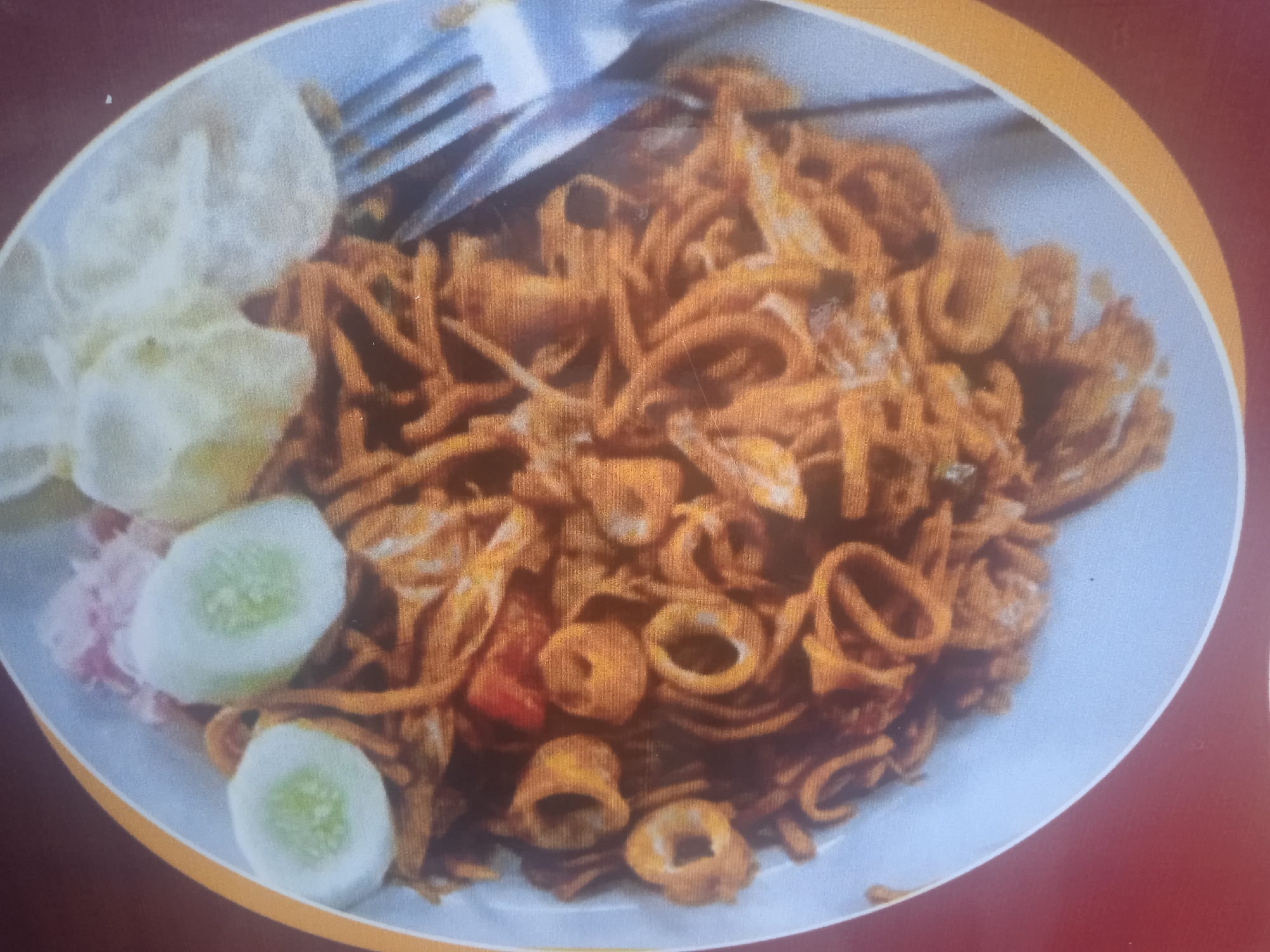 A close-up, overhead shot shows a plate of mie goreng, or fried noodles, garnished with sliced cucumbers and a side of what appears to be crackers and pickled vegetables. The noodles are dark brown and coiled, interspersed with visible rings of squid. A fork is partially visible, resting on the noodles. The plate is white and sits on a dark red surface with a yellow border. The image depicts a food item that might be found at a street food stall or restaurant in Peurupok, Indonesia, a coastal village known for its fresh seafood, making the inclusion of squid a likely culinary element.