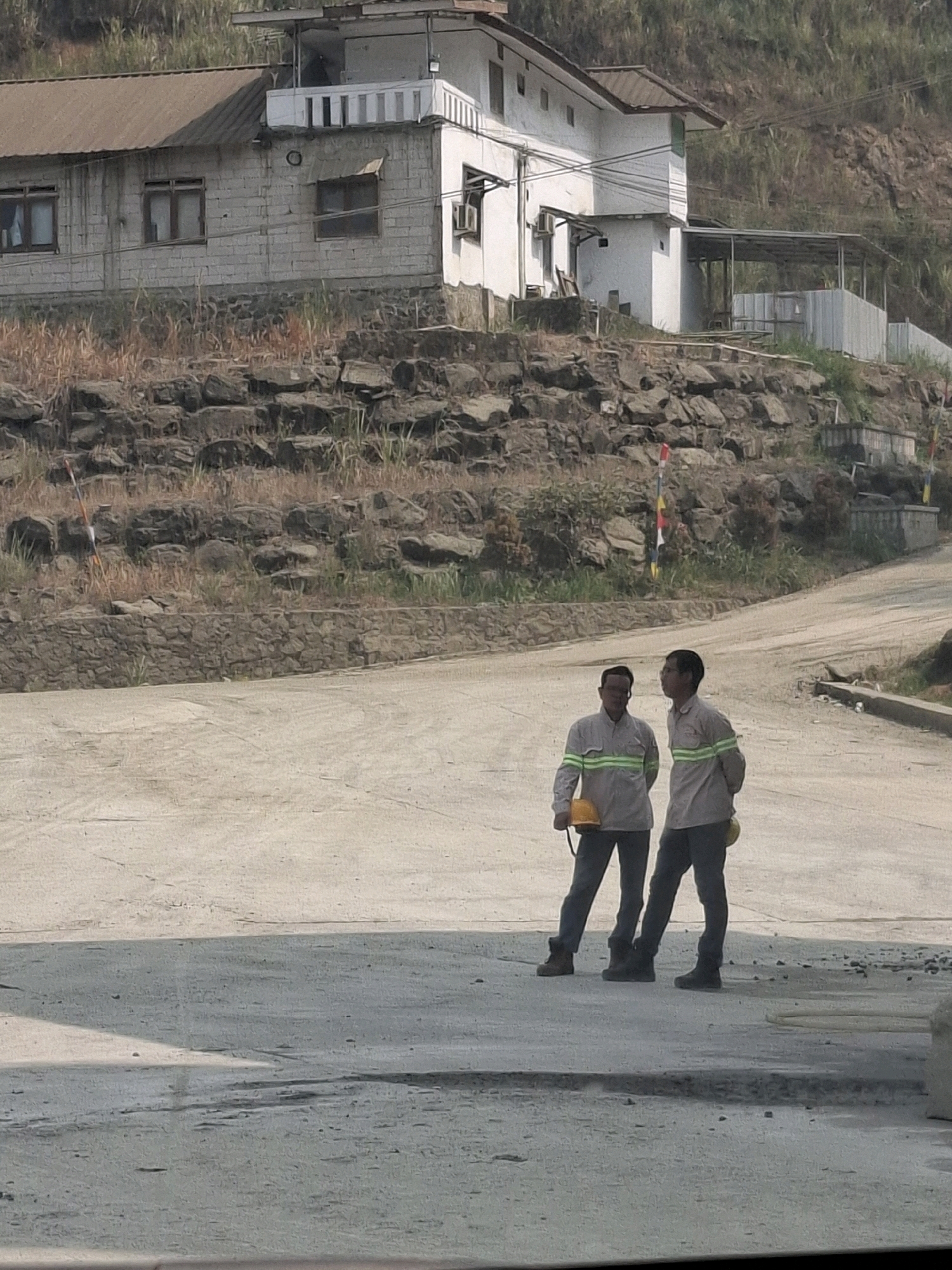 In Cijangkar, Indonesia, two men in work uniforms and safety vests stand on a dusty, unpaved road near a construction site. They are both wearing dark pants and boots, and one holds a yellow hard hat. Behind them, a sloped hill is covered in rocks and dry vegetation. On the hillside, a multi-story building with a white facade and a brown roof is visible. The building has several windows and a balcony with white railings. The overall scene suggests an industrial or construction environment in a hilly, possibly rural, area.