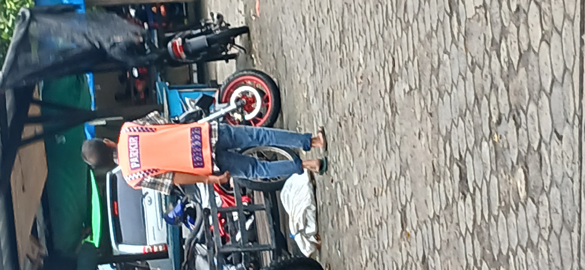 A person wearing a high-visibility orange vest with the word "PARKIR" (Indonesian for "parking") on the back is standing in what appears to be a parking area. The person is wearing a plaid shirt, blue jeans, and flip-flops. The background shows a white vehicle and several motorcycles. The ground is paved with interlocking stones. The scene is likely a public space in Tanjung Balai Karimun, Indonesia, possibly a parking lot or a street with parking.